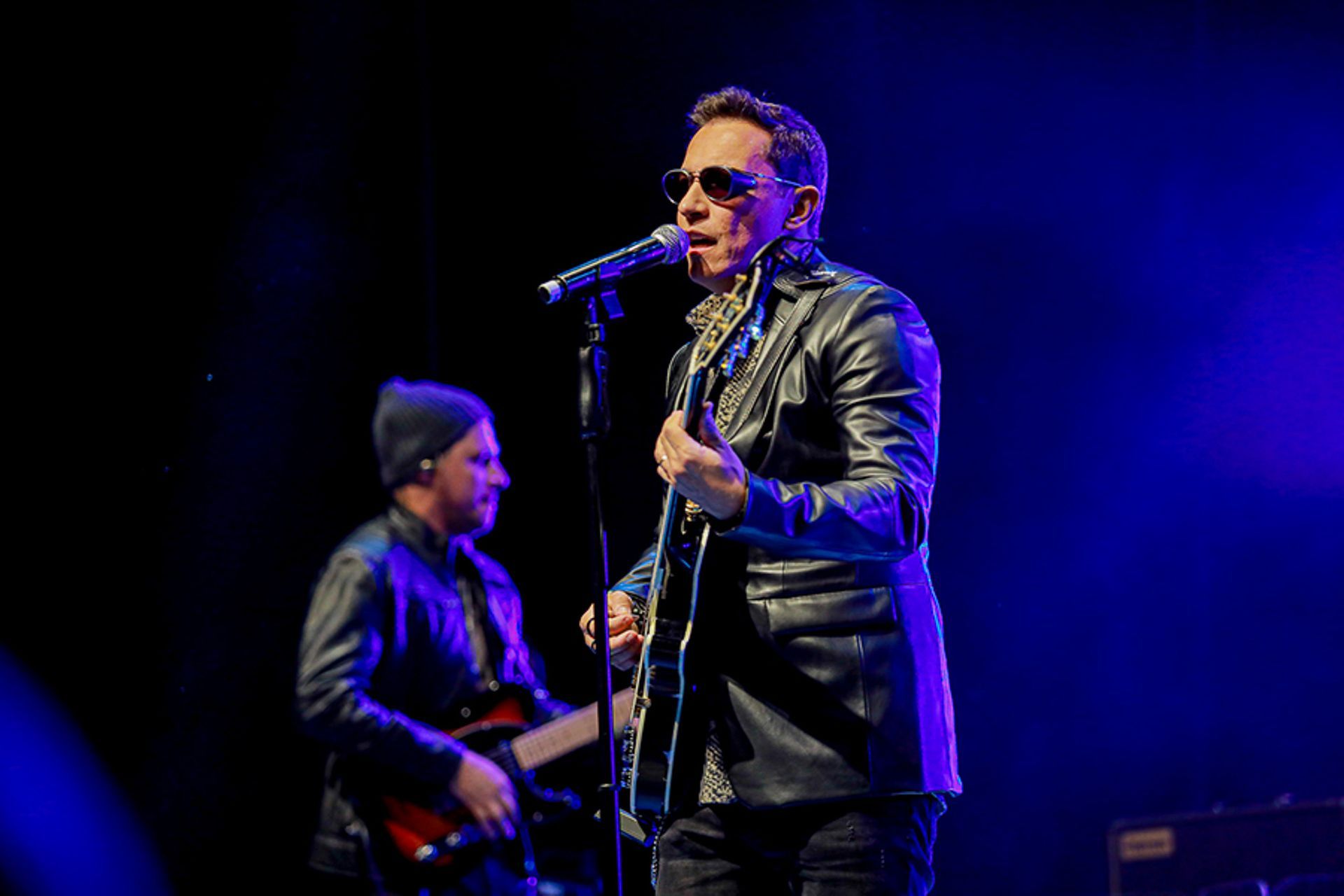 Um artista de jaqueta de couro e óculos escuros canta em um microfone no palco, com um músico tocando violão atrás dele.