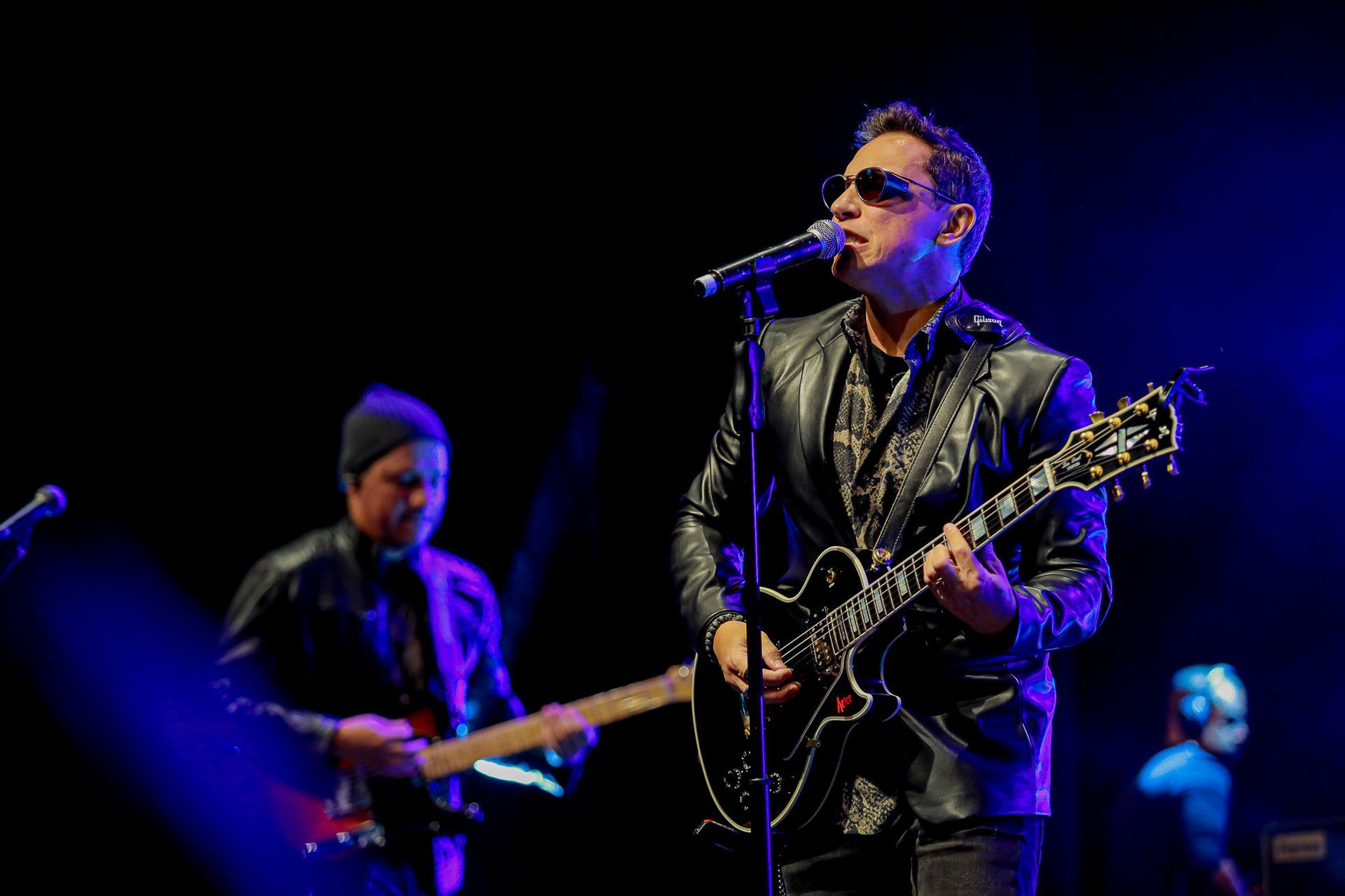 Um cantor de óculos escuros e jaqueta de couro preta se apresenta com uma guitarra em um palco de shows com pouca luz.