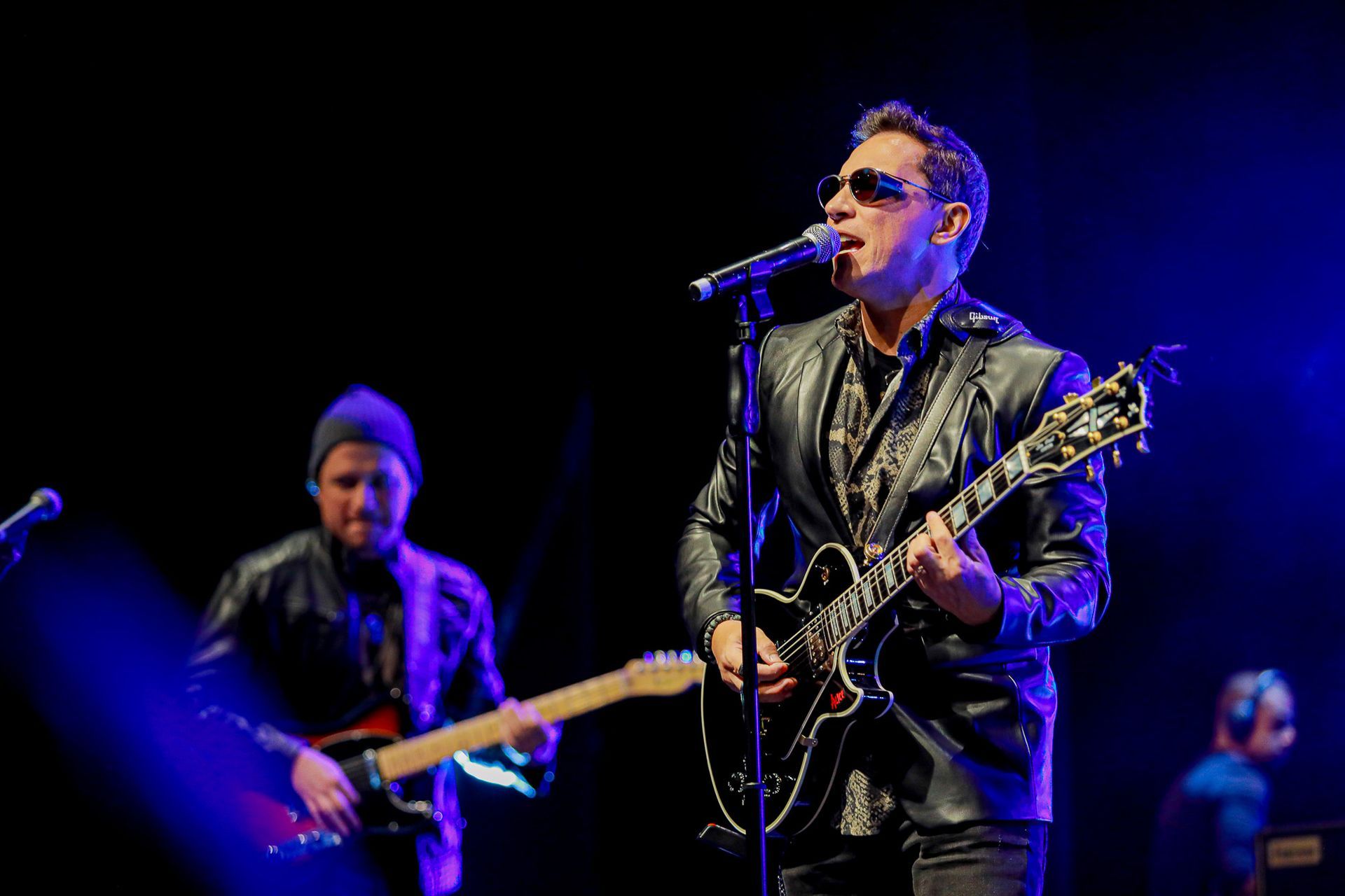 Um músico canta ao microfone enquanto toca guitarra elétrica no palco, com um membro da banda tocando guitarra atrás dele.