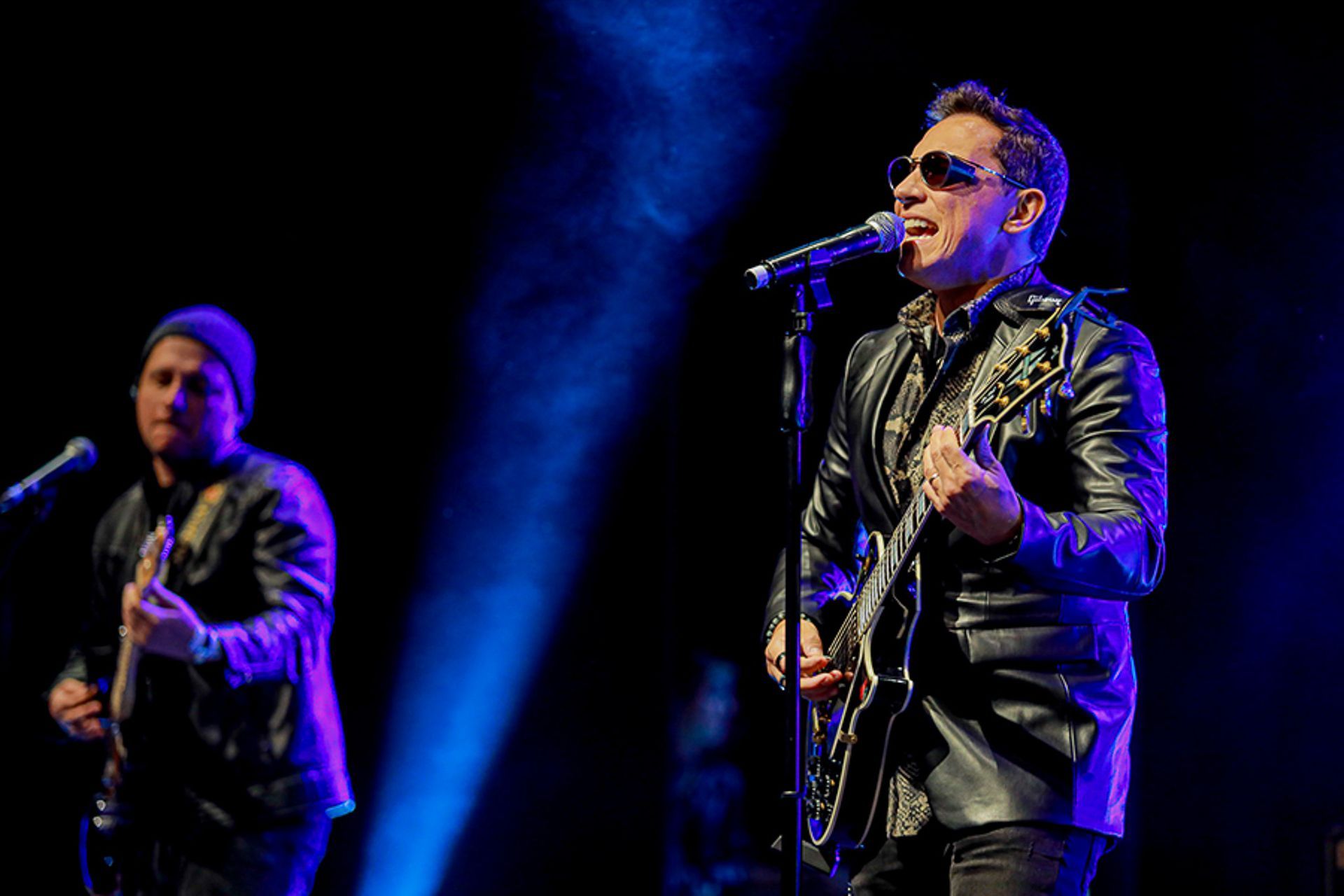 Dois músicos se apresentam no palco sob holofotes azuis; o vocalista usa óculos escuros e toca guitarra elétrica.