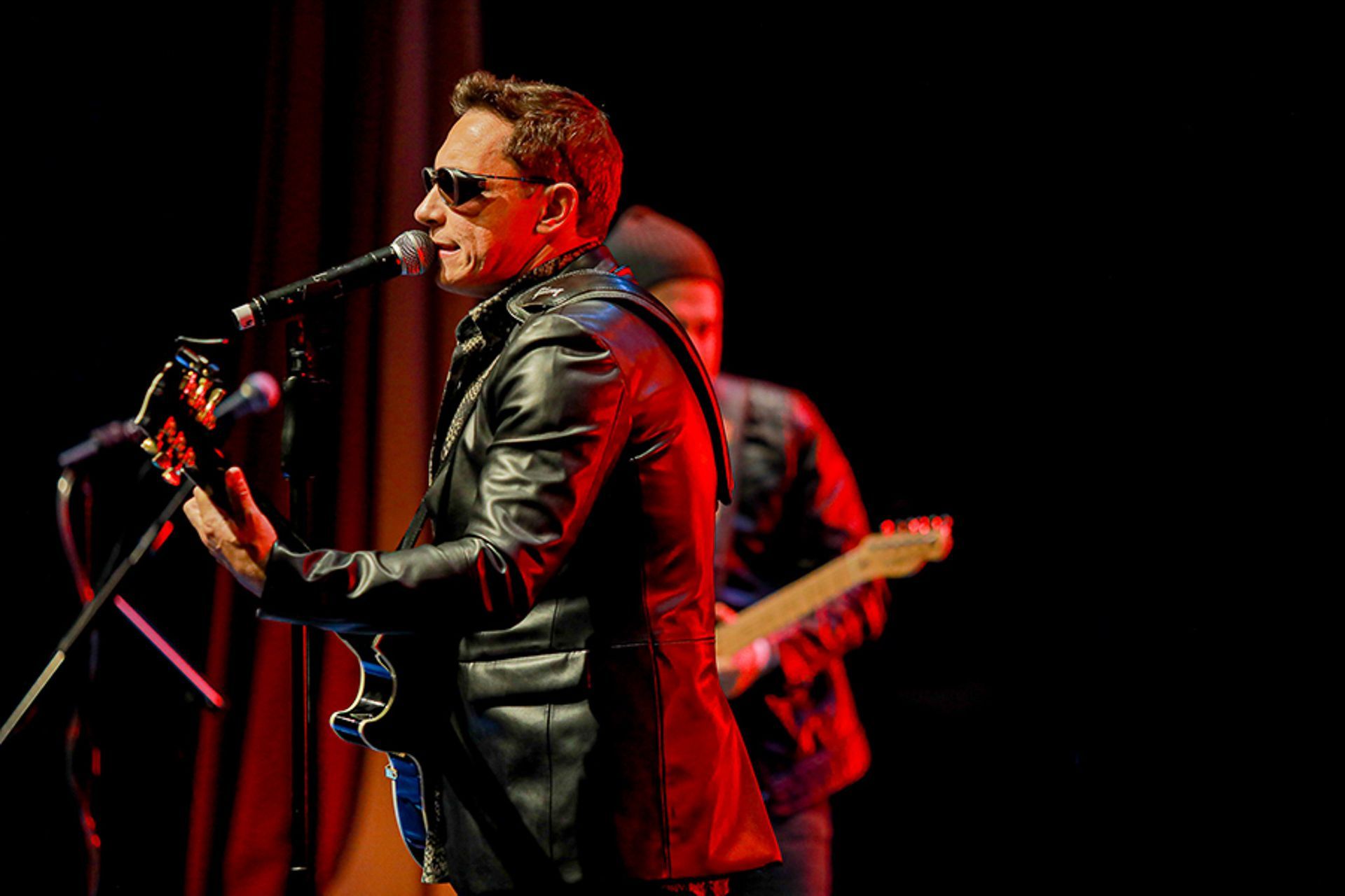Um músico de jaqueta de couro e óculos escuros toca guitarra elétrica enquanto canta ao microfone no palco.