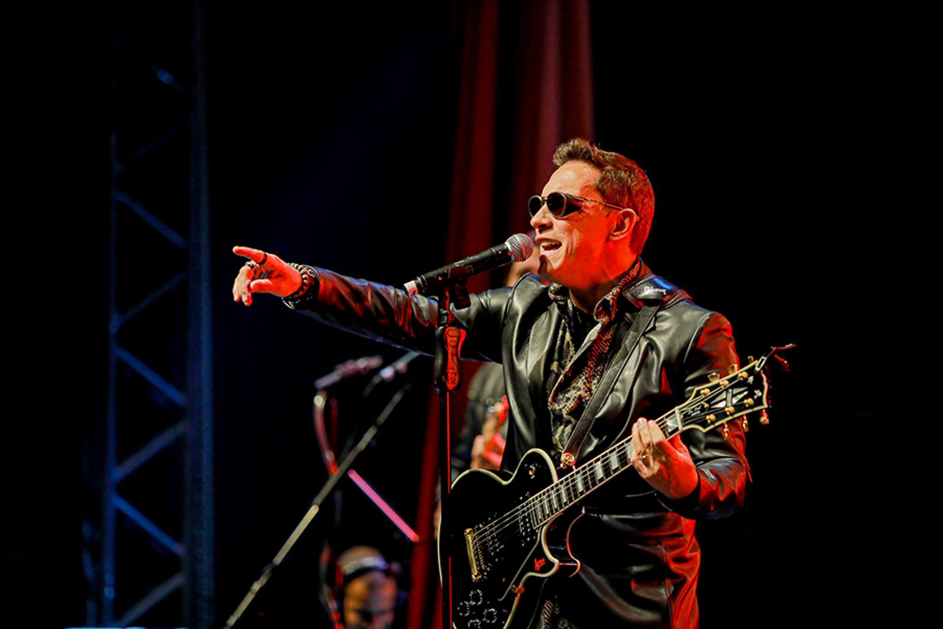 Um cantor de jaqueta preta e óculos escuros aponta enquanto toca guitarra elétrica em um microfone no palco.