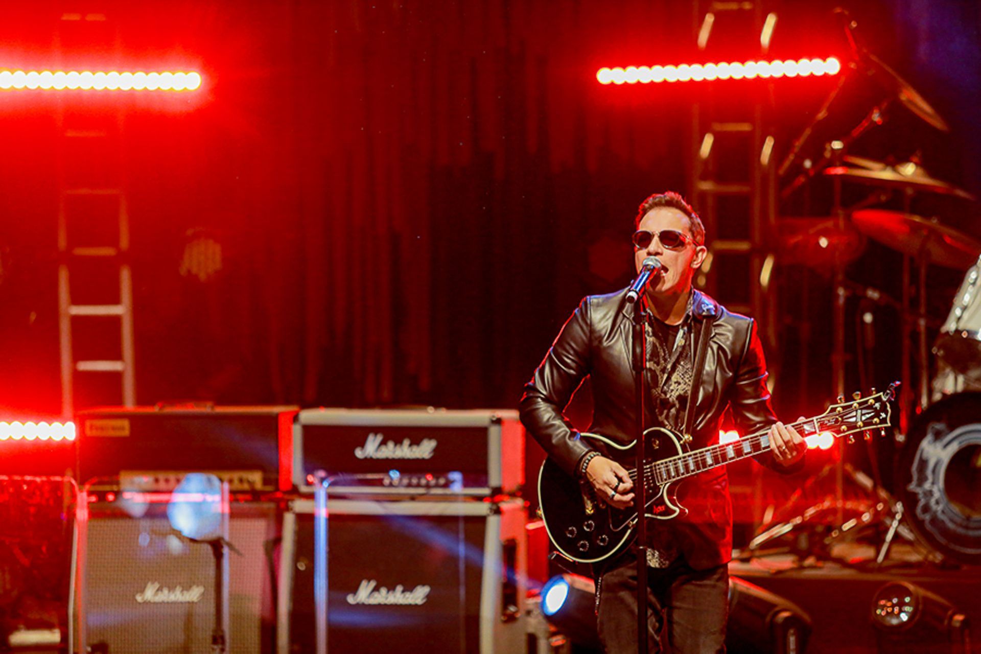 Um músico de óculos escuros toca guitarra elétrica enquanto canta em um microfone em um palco com amplificadores Marshall.