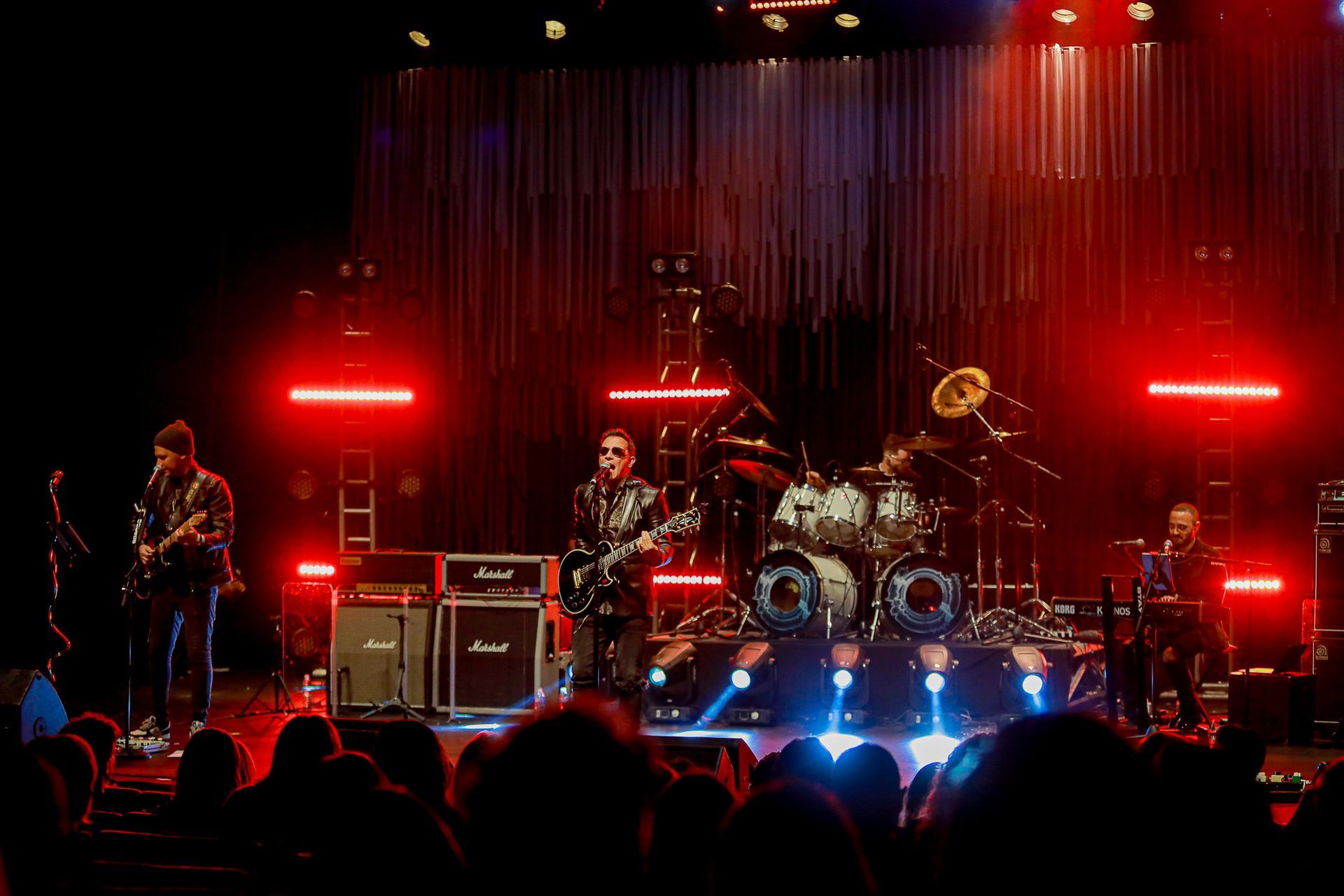 Uma banda se apresenta em um palco com iluminação baixa e luz, composta por um guitarrista, um vocalista e um baterista.