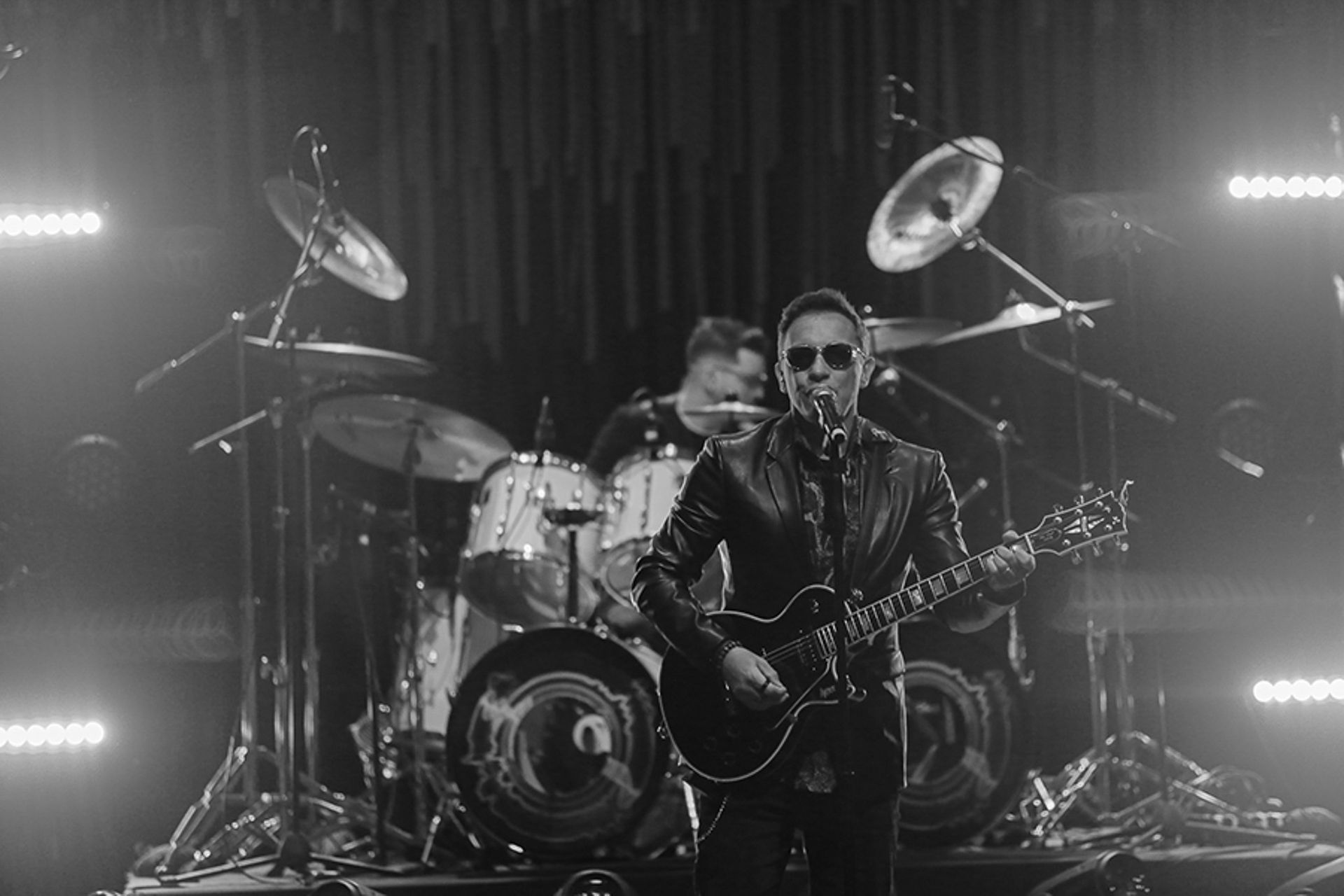 Um músico usando óculos escuros toca guitarra elétrica no palco com um baterista ao fundo, sob a iluminação do palco.