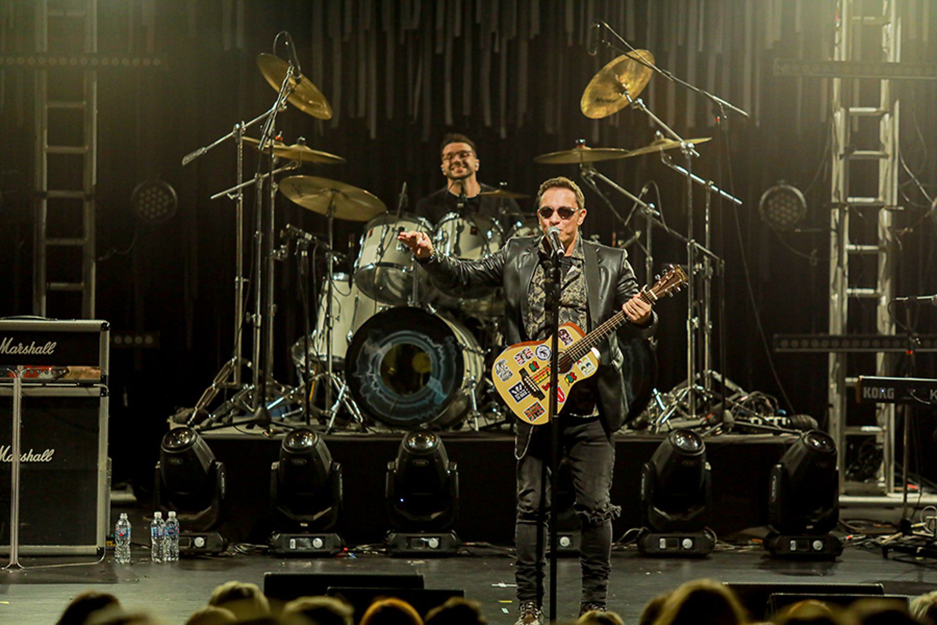 Um cantor toca violão em um palco com um baterista atrás dele, ambos sob holofotes aconchegantes em uma casa de shows.