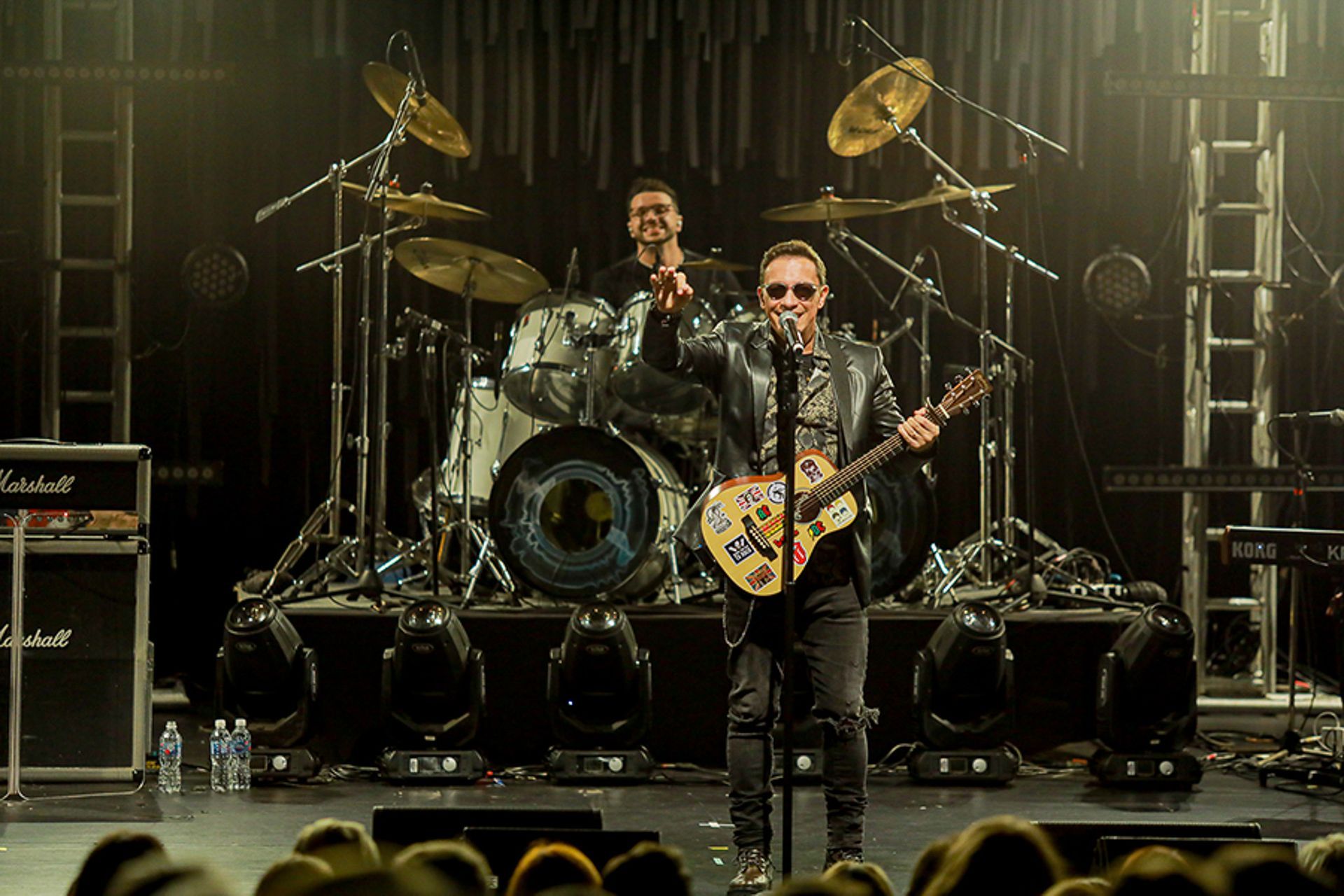 Um músico de jaqueta de couro toca um violão coberto de adesivos no palco, com um baterista tocando atrás dele.