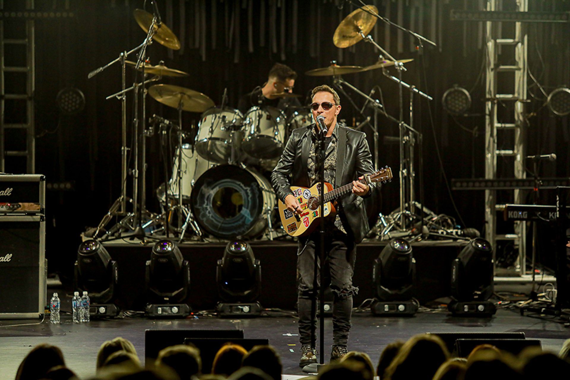 Um músico usando óculos escuros e jaqueta de couro se apresenta com um violão no palco diante de uma plateia.