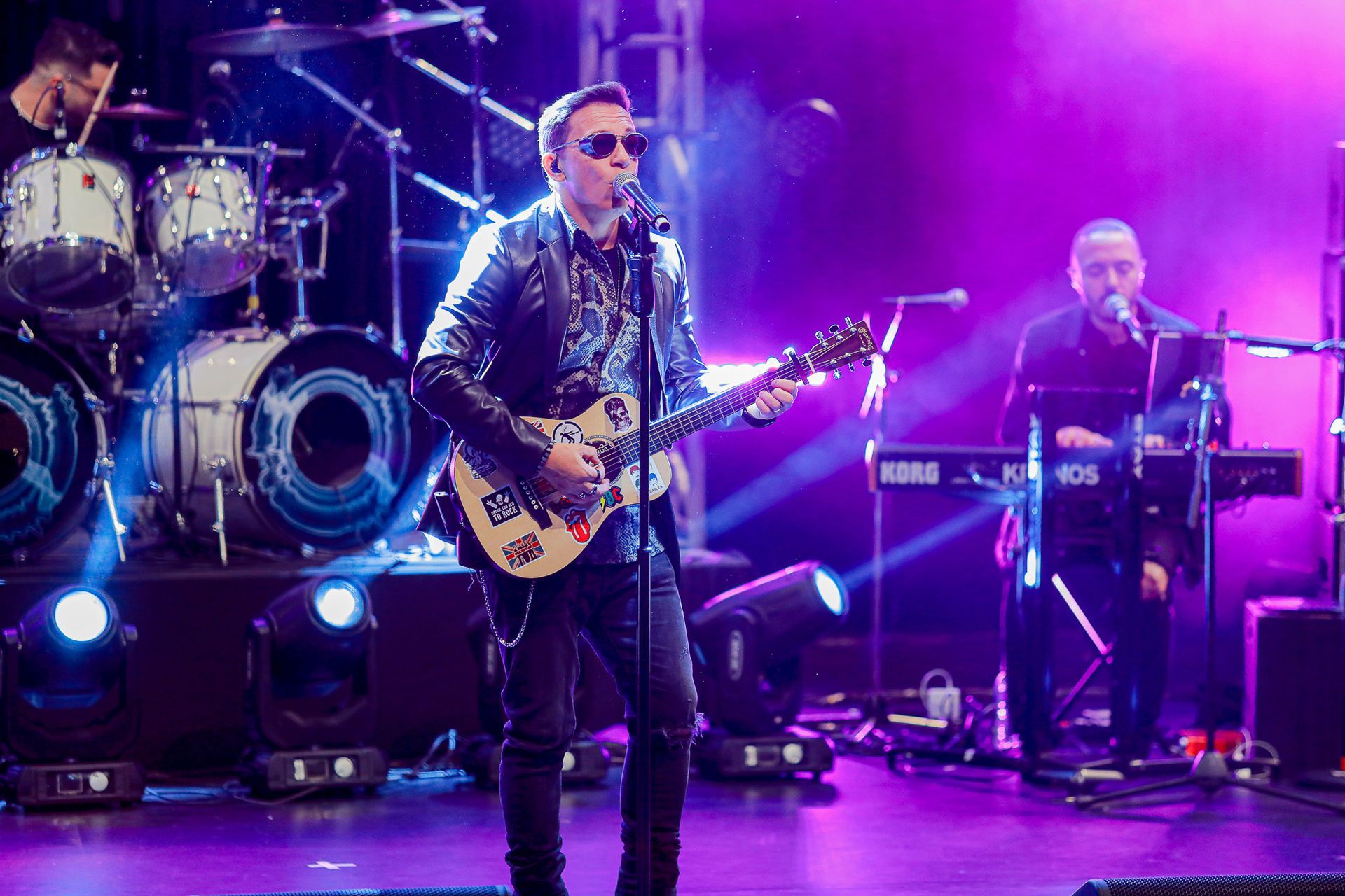 Um artista tocando guitarra elétrica e cantando ao microfone em um palco com iluminação roxa e um tecladista.
