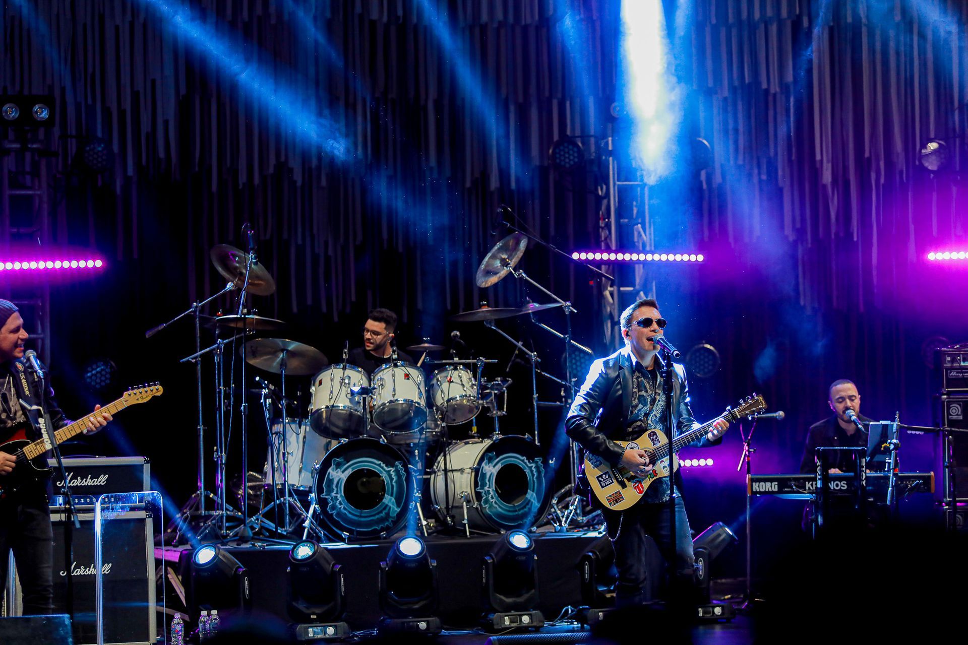 Uma banda de rock ao vivo se apresenta em um palco iluminado, composta por guitarrista, baterista, vocalista e tecladista.