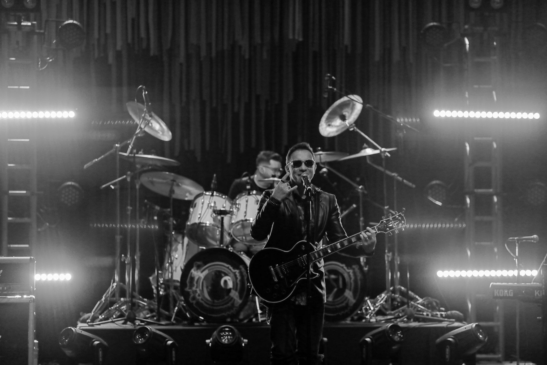 Uma foto em preto e branco de um músico tocando guitarra elétrica no palco, com um baterista visível ao fundo.