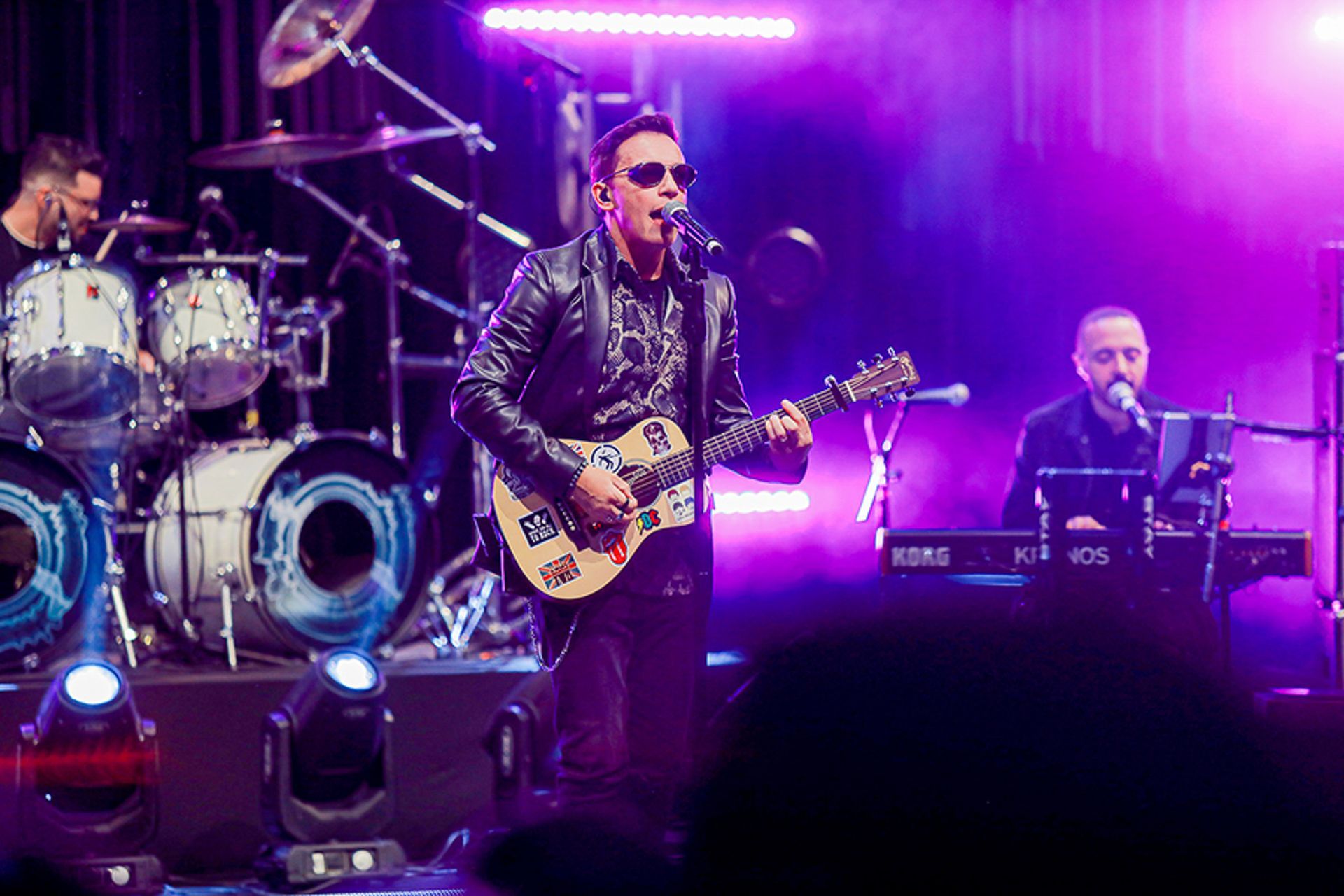 Um músico cantando e tocando violão em um palco iluminado com luzes roxas, acompanhado por um baterista e um tecladista.