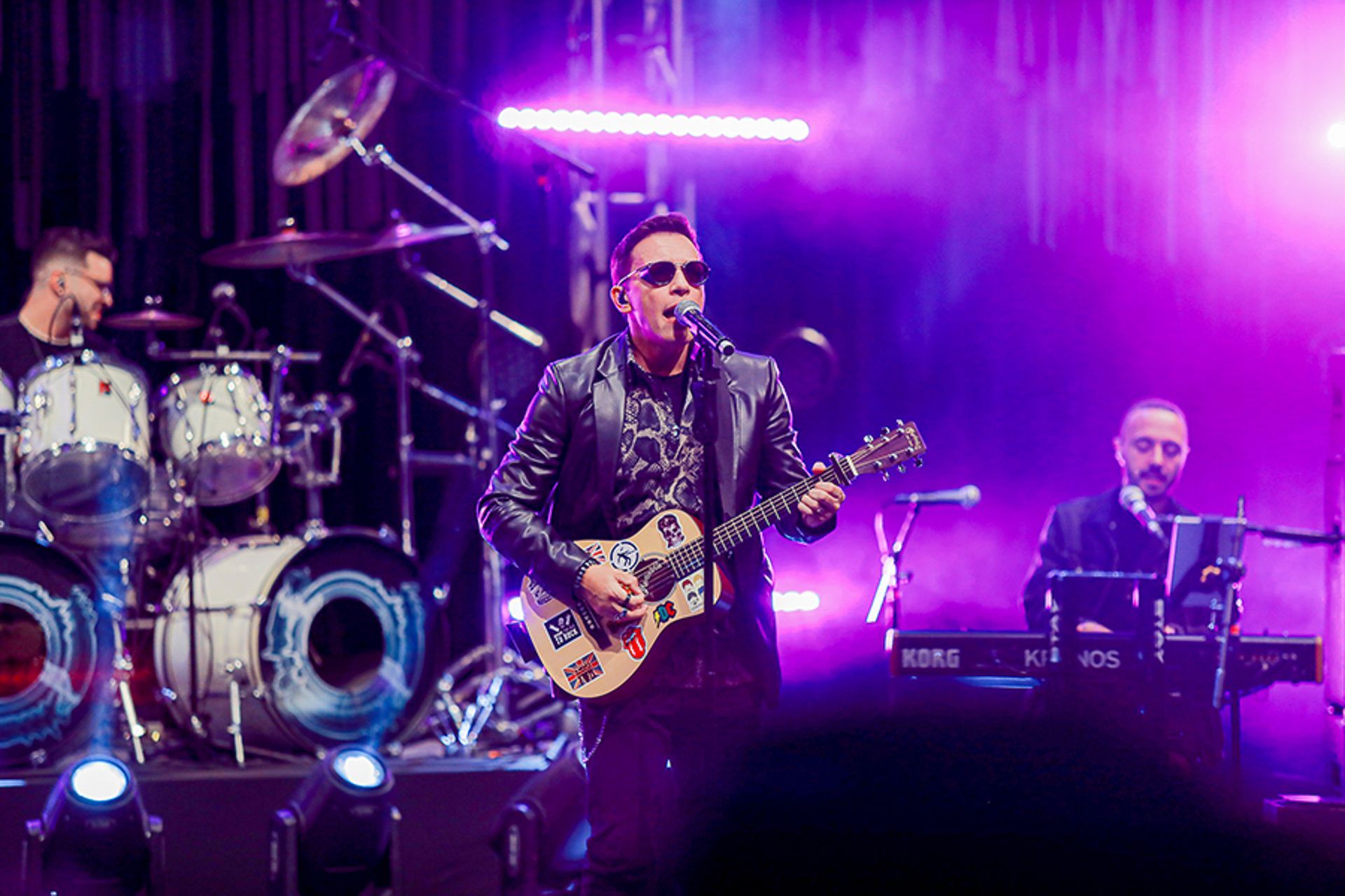 Um músico canta ao microfone enquanto toca violão em um palco iluminado por luzes roxas brilhantes.