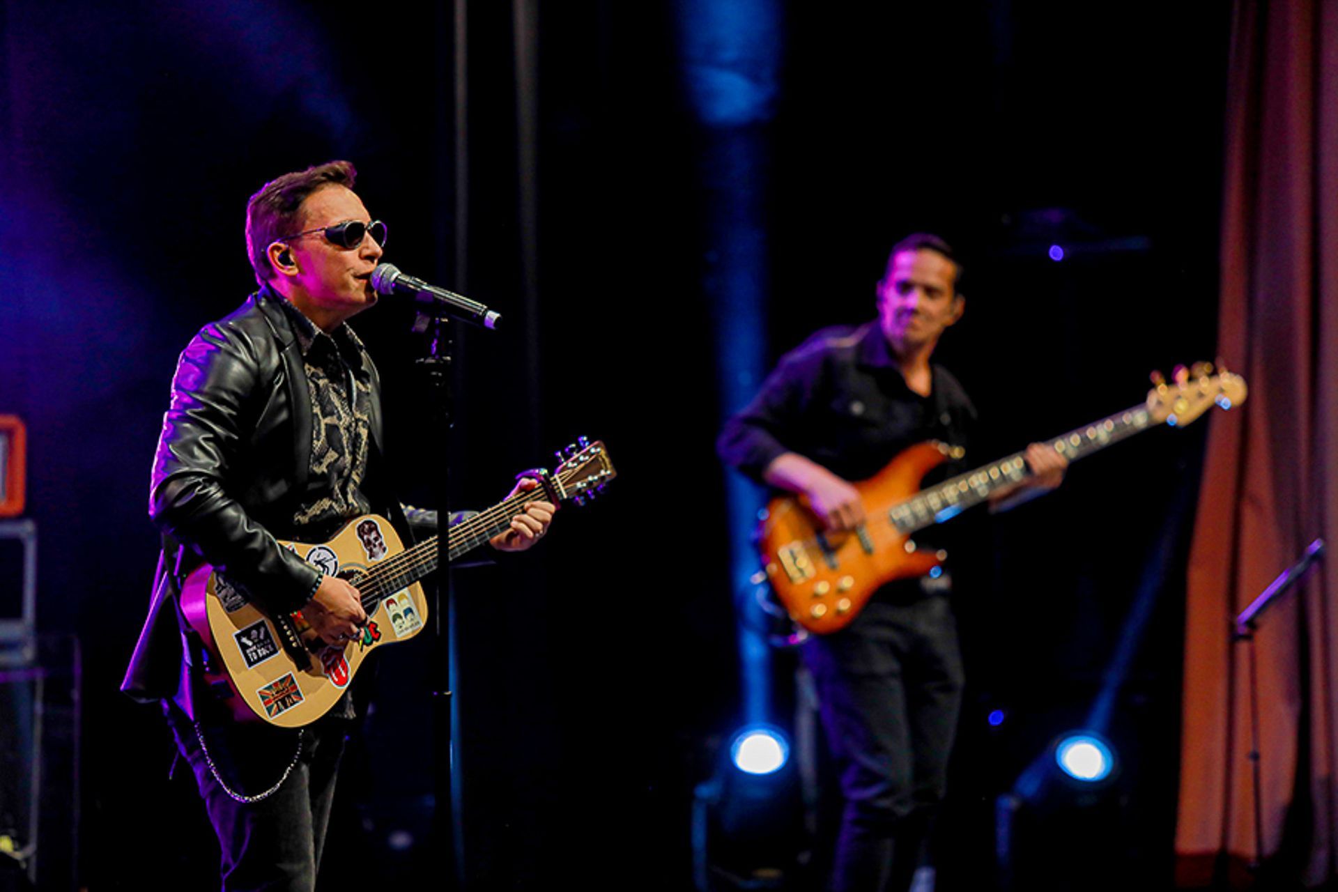 Um artista de jaqueta de couro canta ao microfone enquanto toca guitarra, acompanhado por um baixista em um palco escuro.