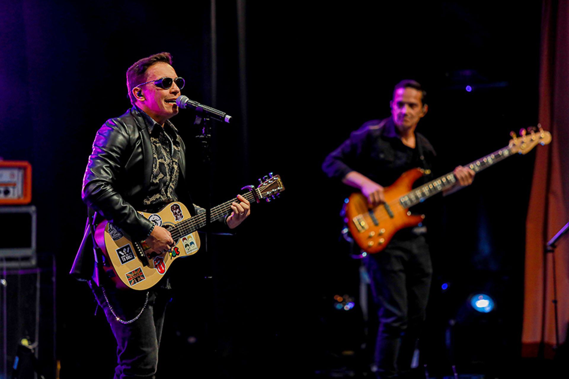 Dois músicos se apresentando em um palco escuro; um vocalista com um violão e um baixista tocando ao fundo.