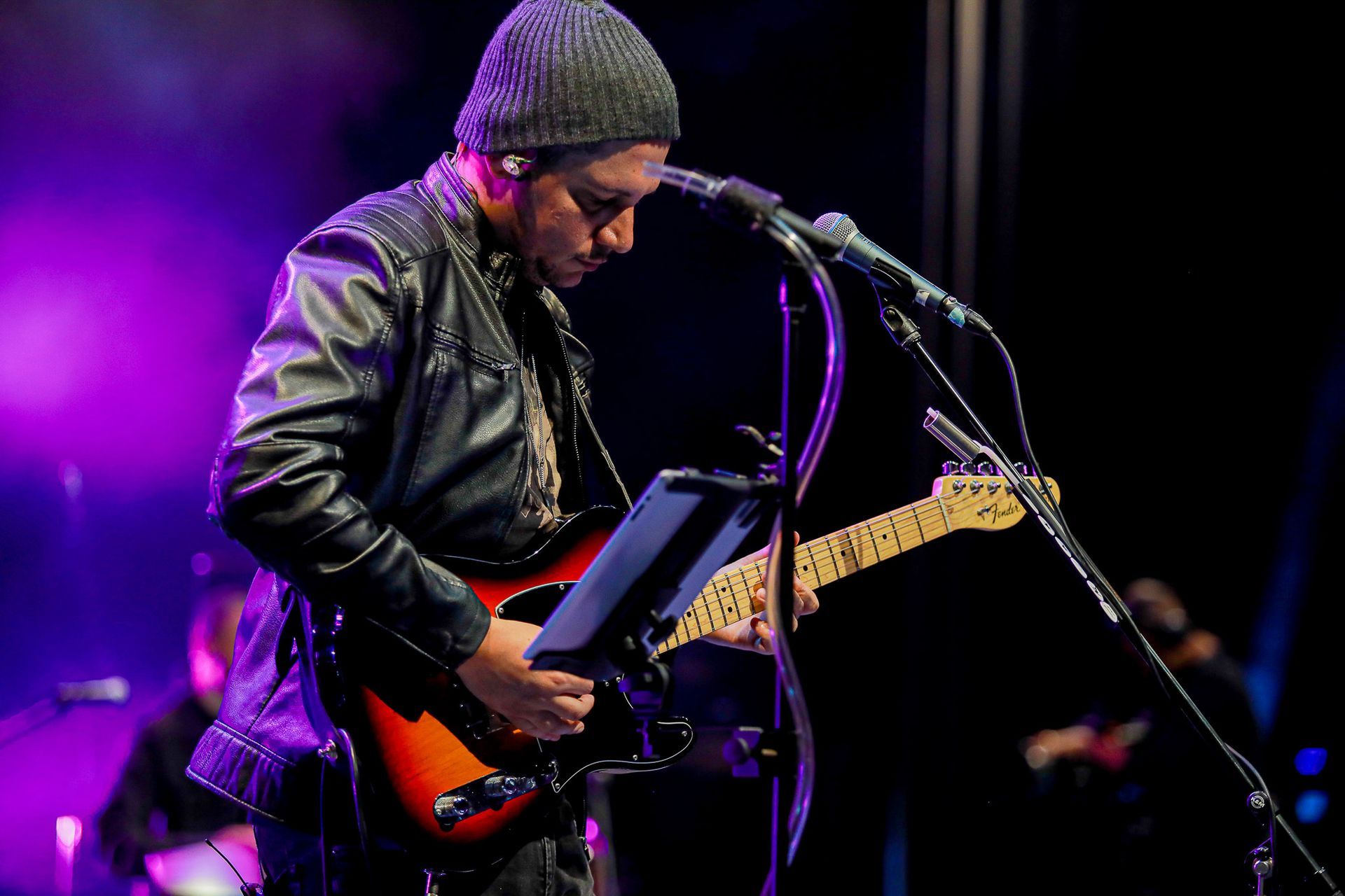 Um músico de gorro e jaqueta de couro toca uma guitarra elétrica sunburst em um palco pouco iluminado com luzes roxas.