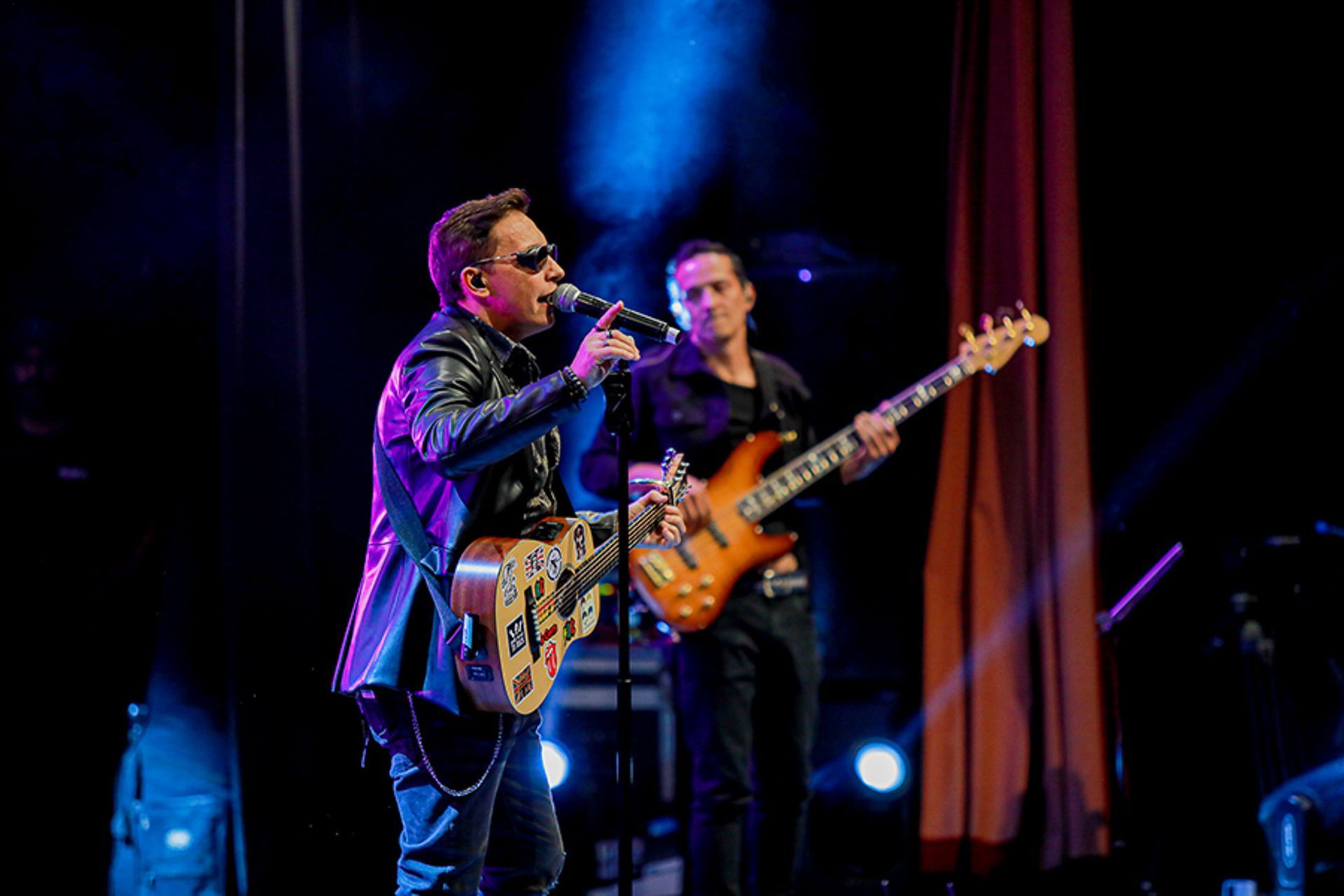 Dois músicos se apresentam em um palco com iluminação azul, um deles cantando ao microfone enquanto toca violão.