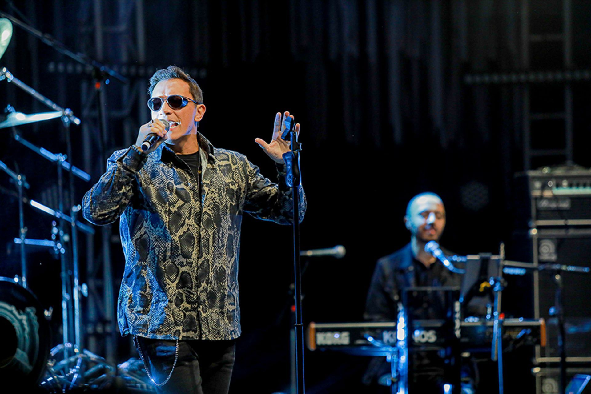 Um cantor vestindo uma camisa estampada de botões e óculos escuros se apresenta ao microfone no palco, perto de um tecladista