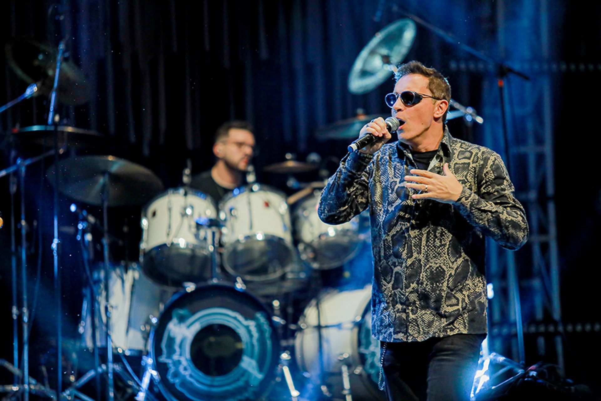O vocalista, usando óculos escuros e uma camisa estampada, se apresenta ao microfone no palco, com um baterista visível.