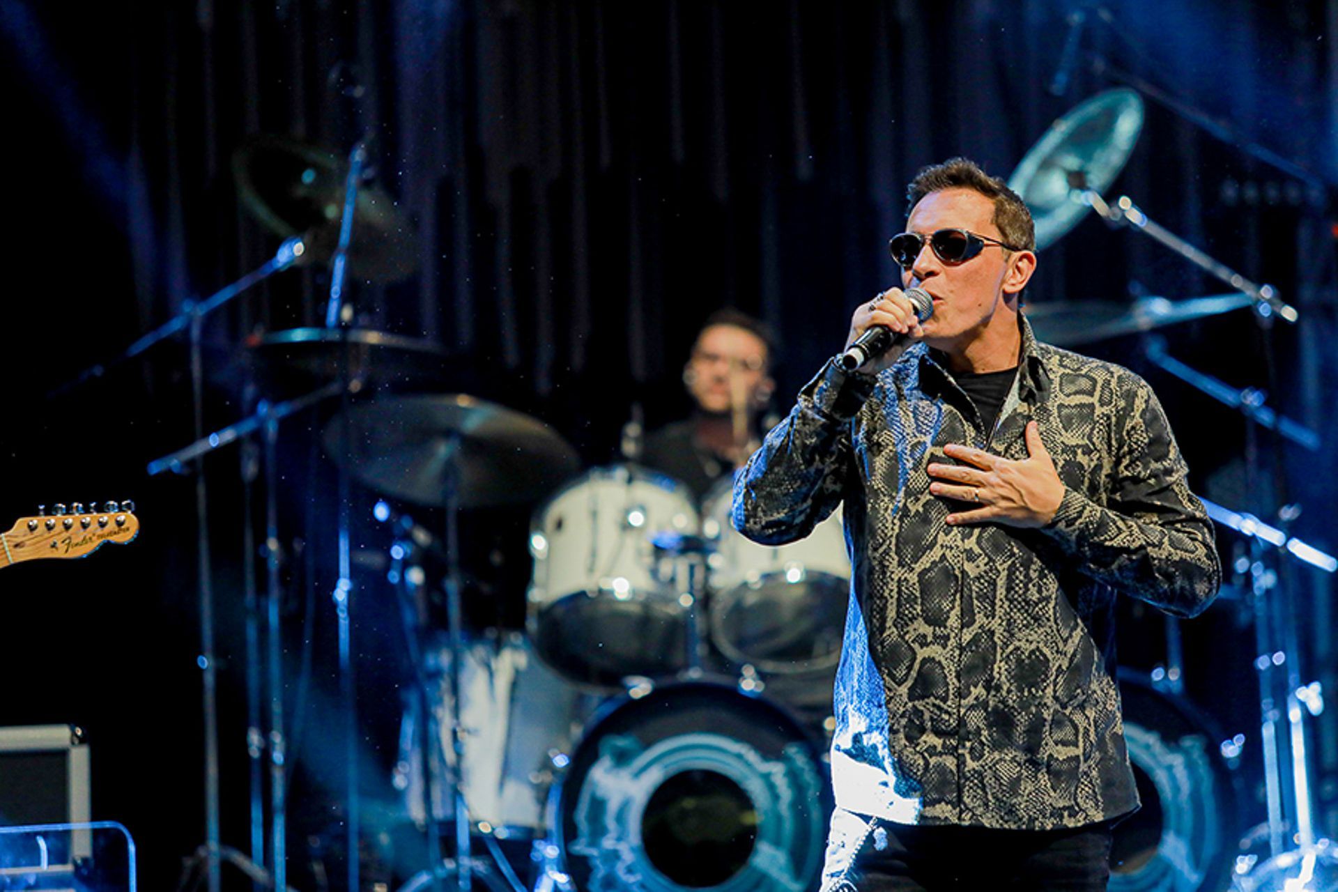 O vocalista, vestindo uma camisa estampada de botões e óculos escuros, canta ao microfone no palco, em frente a um baterista.