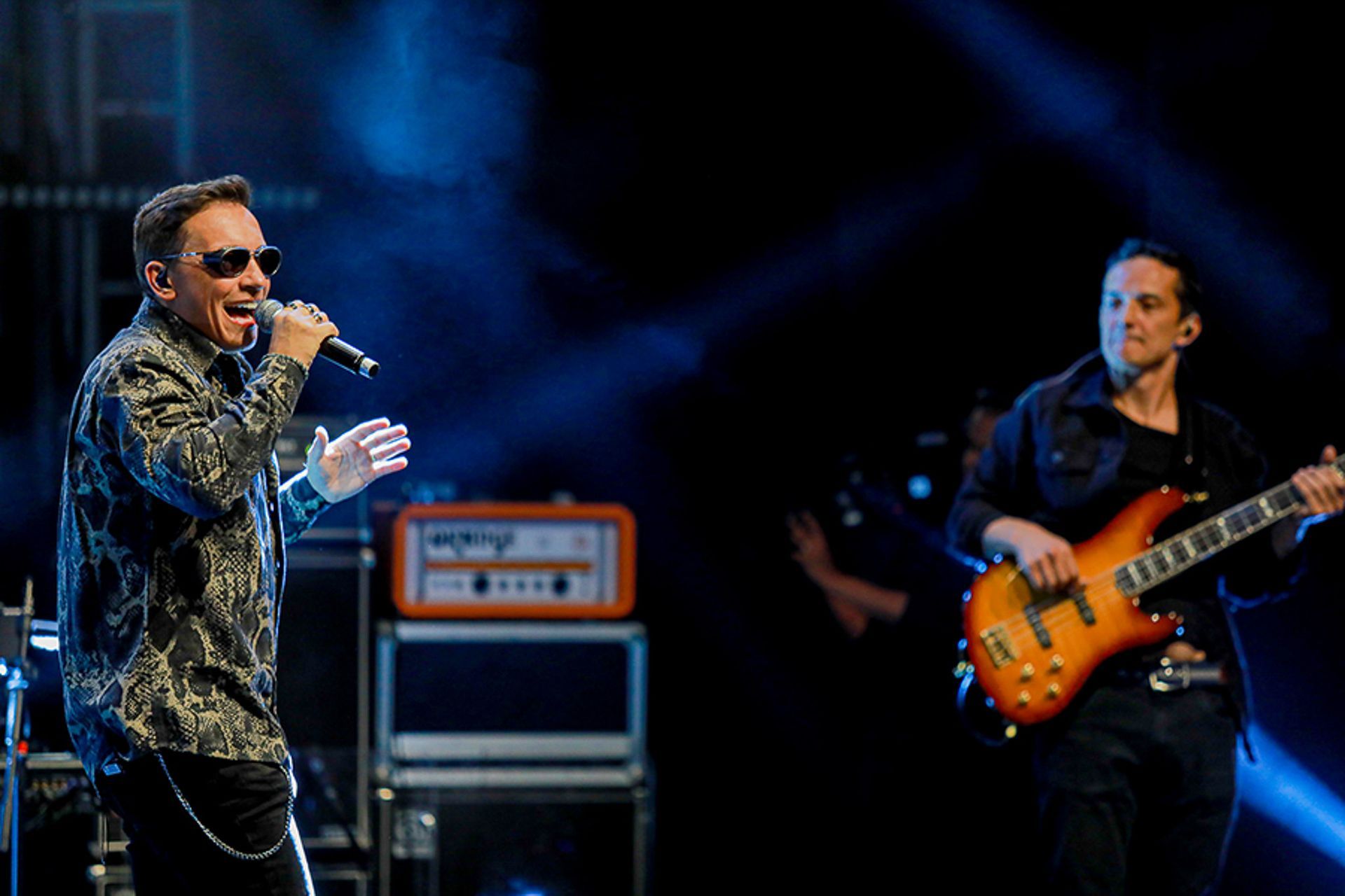 O vocalista, vestindo uma camisa estampada, canta em um microfone ao lado de um baixista que toca um baixo no palco.