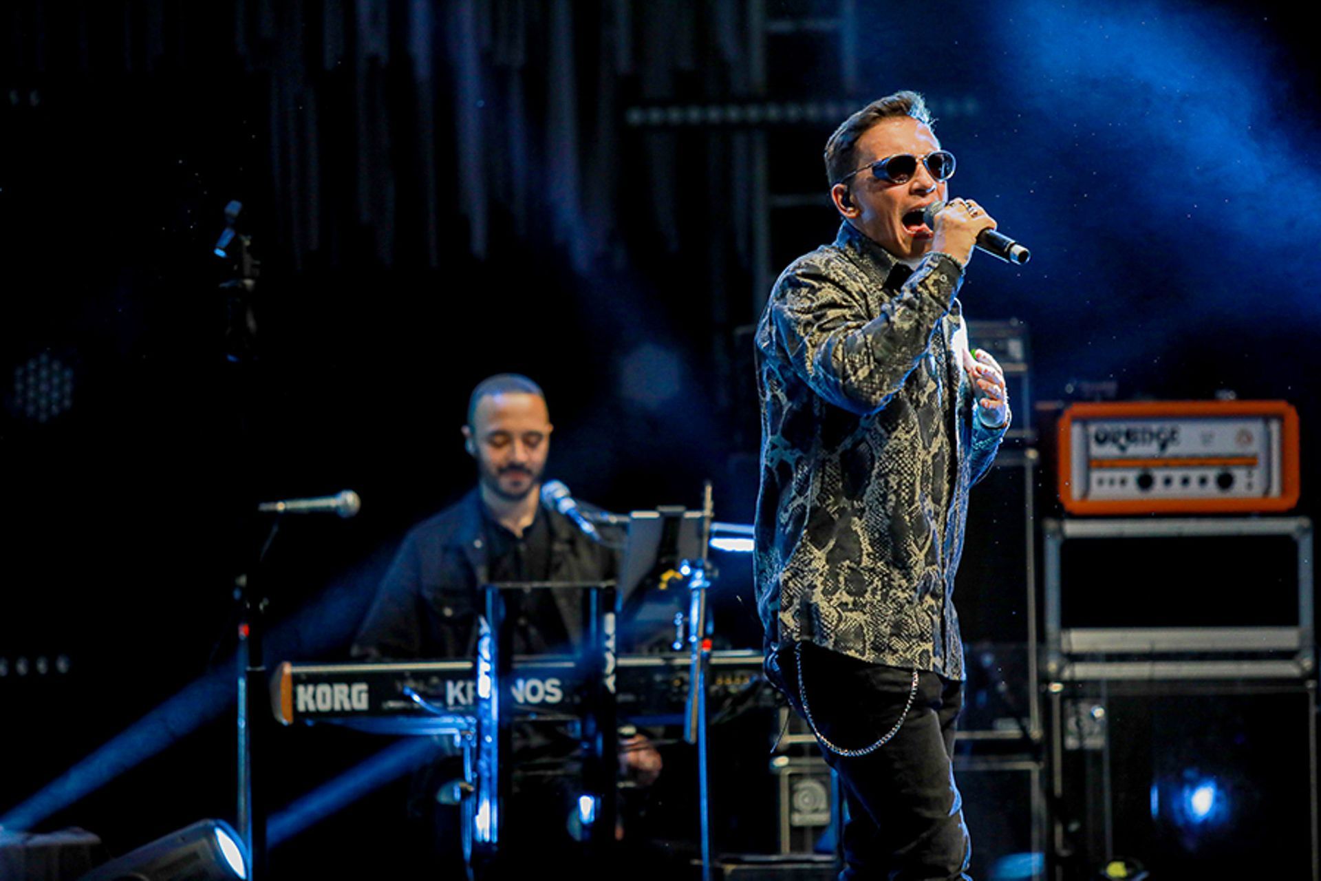 Um cantor com uma camisa estampada se apresenta ao microfone no palco, enquanto um tecladista toca ao fundo.