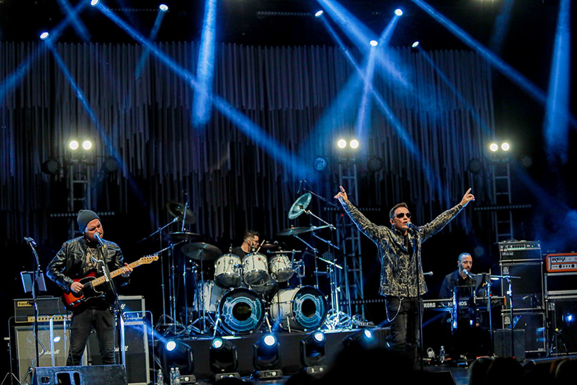 Uma banda se apresenta em um palco sob holofotes azuis brilhantes, com o vocalista principal, em direção às luzes.