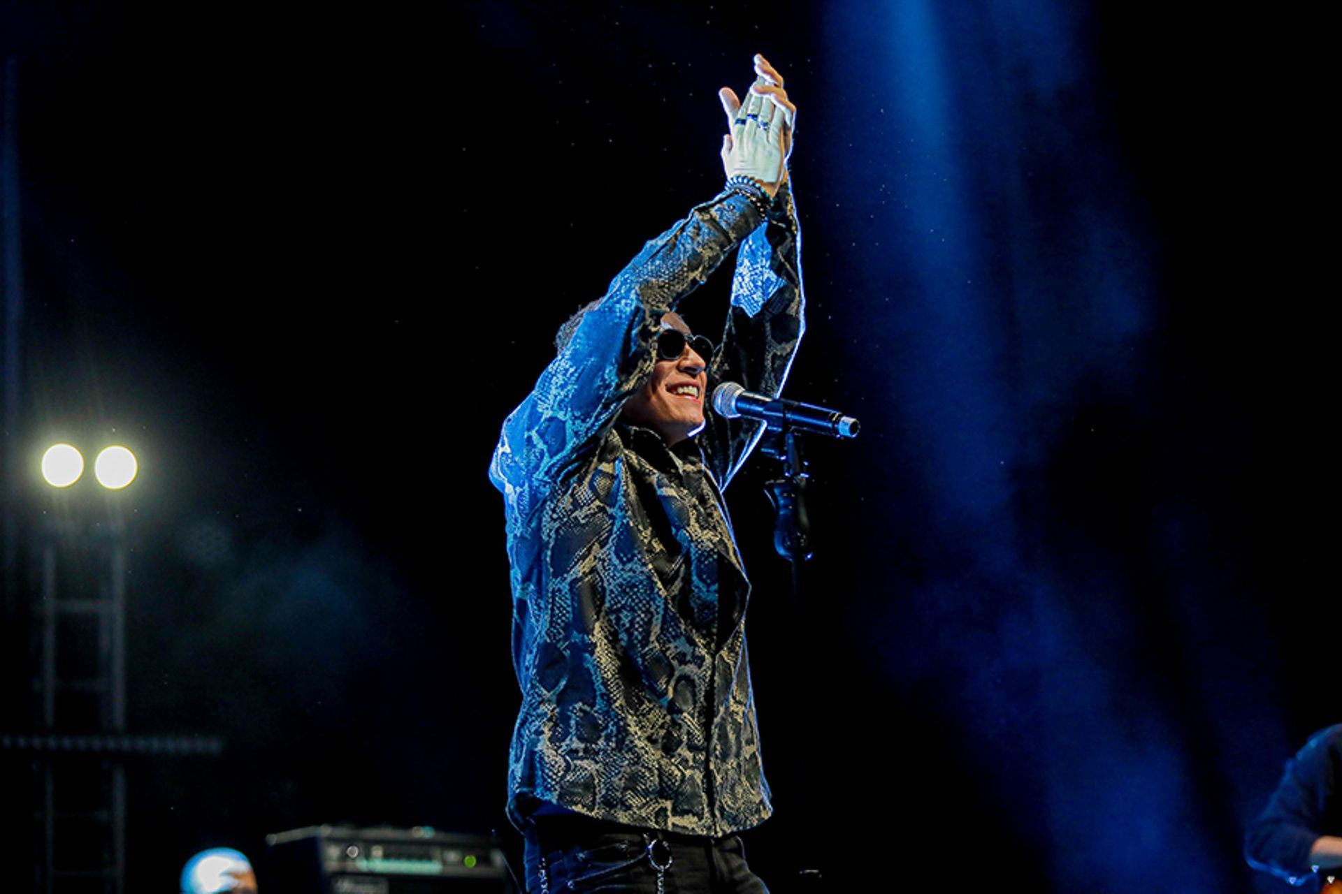 Um artista vestindo uma camisa estampada e óculos escuros no palco, batendo palmas com os braços erguidos sob a iluminação.