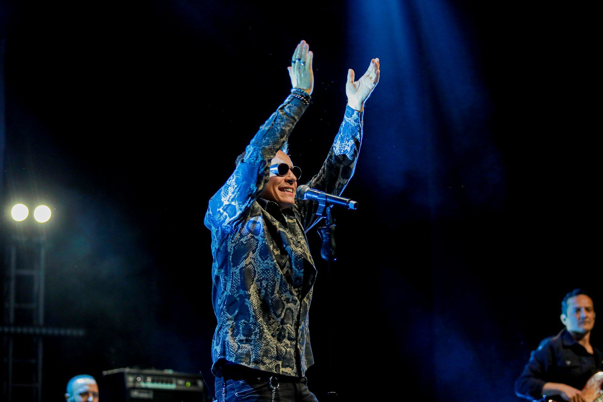 Um artista vestindo uma jaqueta azul estampada e óculos escuros bate palmas no palco sob a iluminação azul de um show.