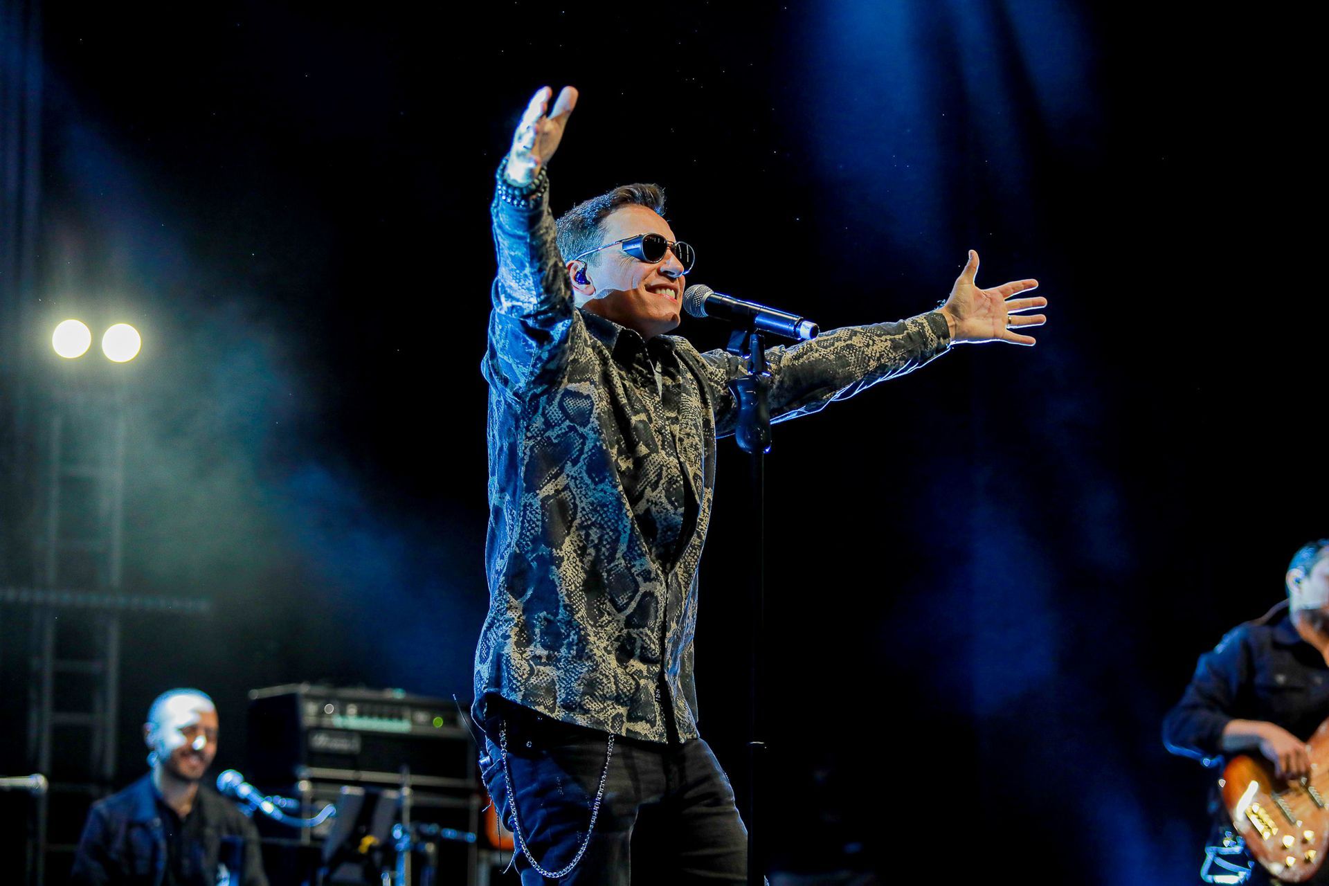 Um artista de óculos escuros e camisa estampada canta em um microfone com os braços estendidos no palco.