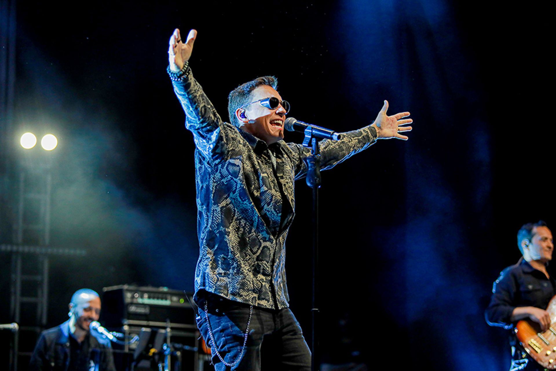 Uma cantor vestindo uma camisa estampada de botões e óculos escuros se apresenta no palco, iluminada pelas luzes do palco.