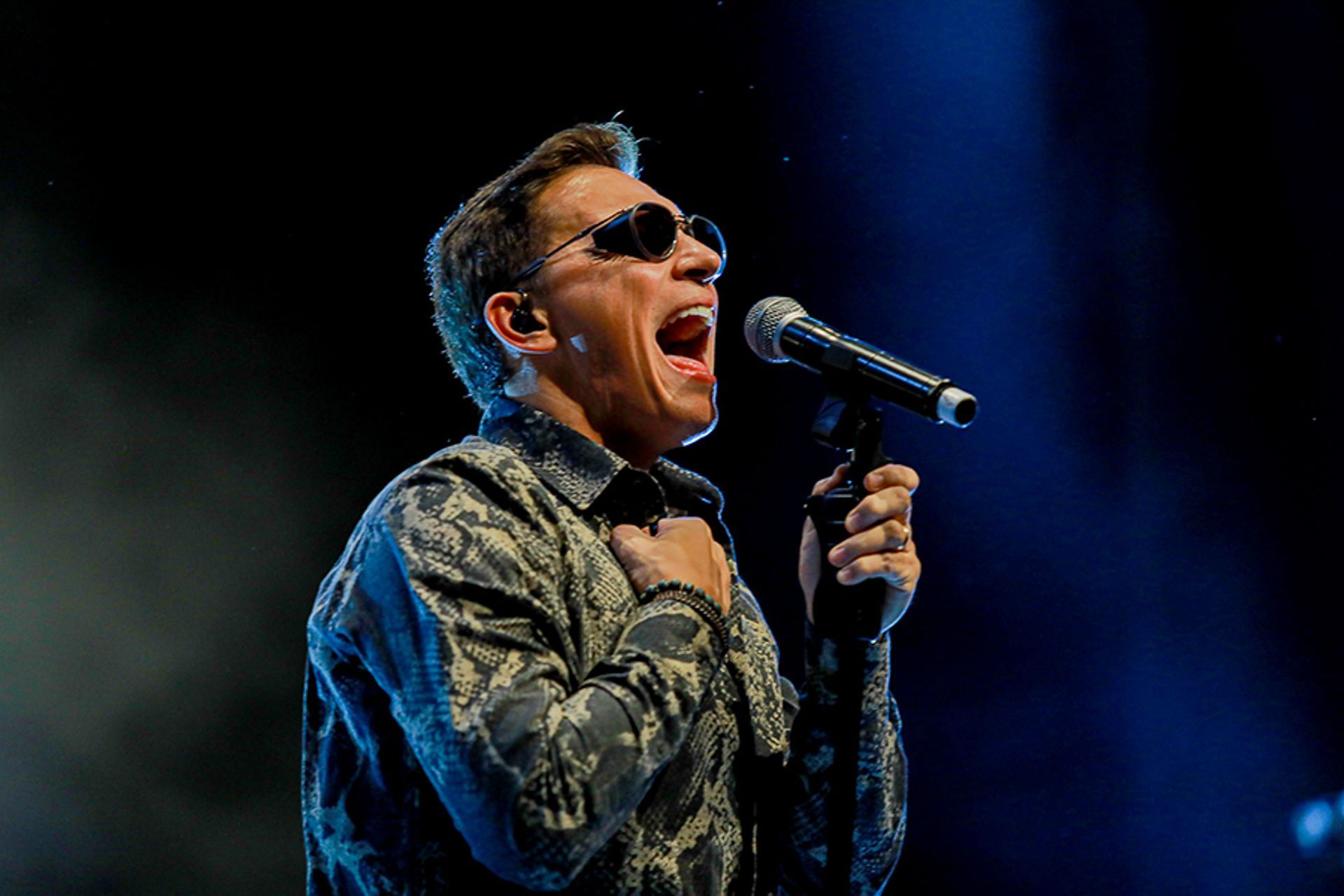 Uma pessoa vestindo uma camisa estampada e óculos escuros cantando com paixão em um microfone em um palco pouco iluminado.