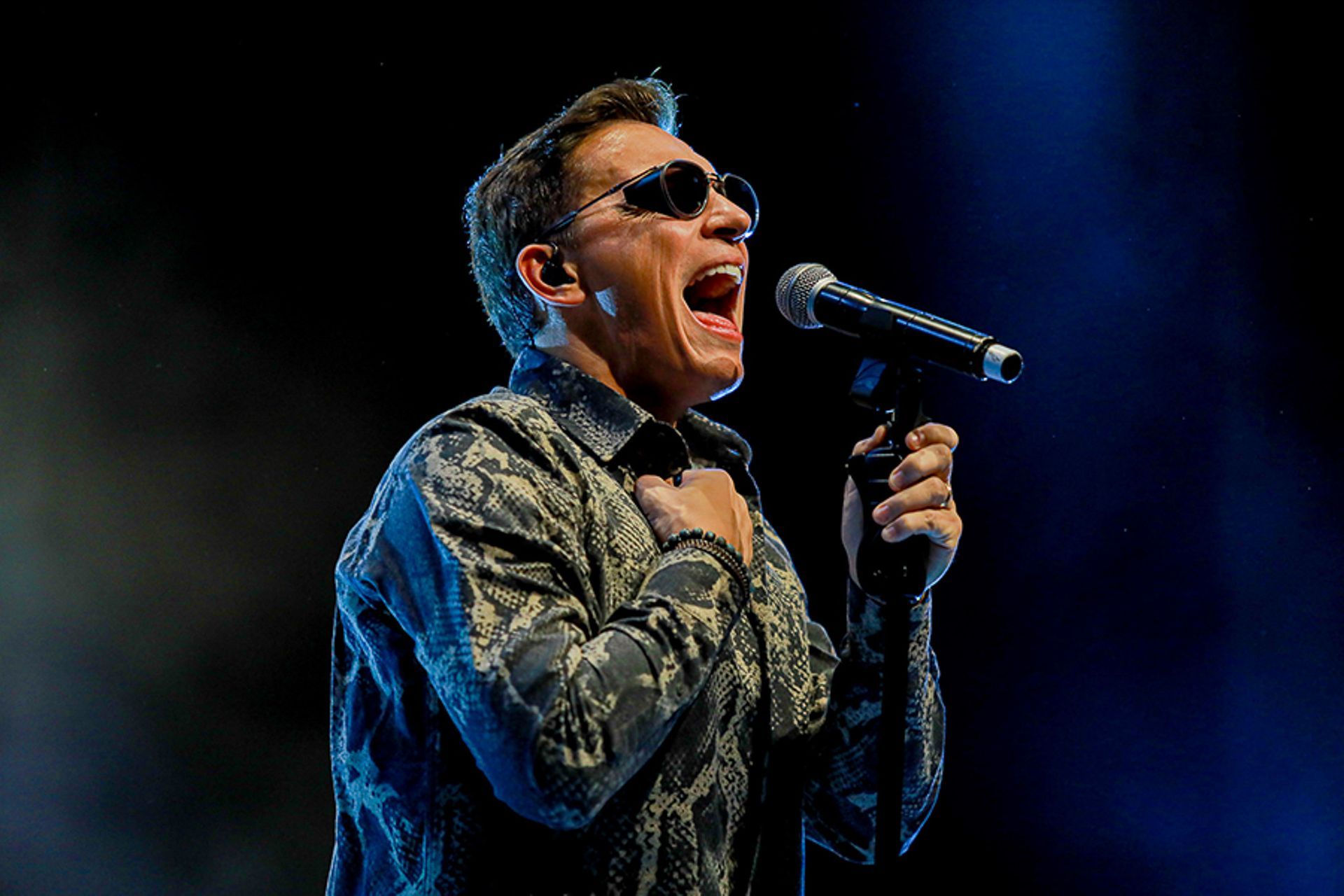 Um artista cantando ao microfone, usando óculos escuros e uma camisa estampada contra um fundo escuro com iluminação azul.
