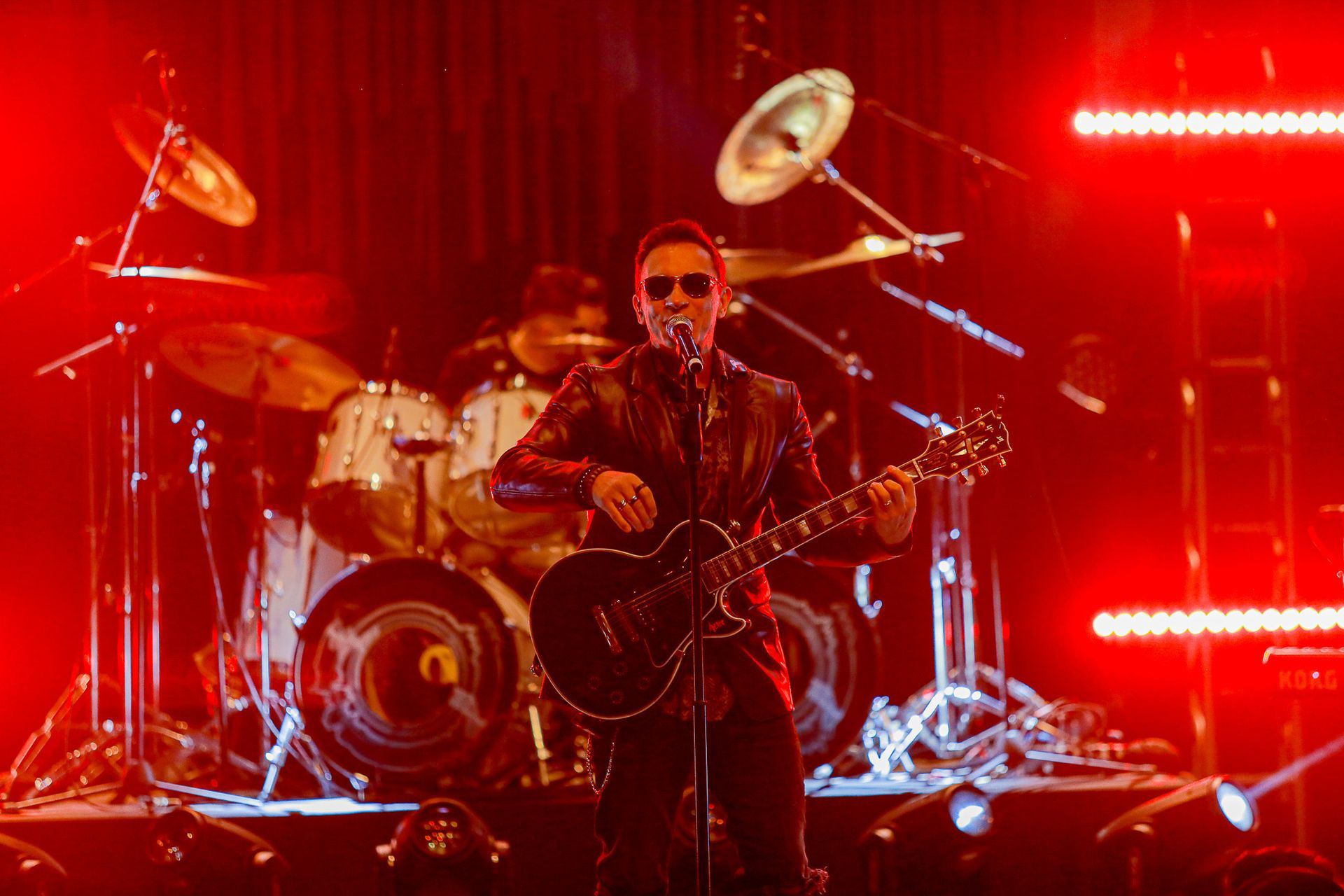 Um cantor de óculos escuros toca guitarra elétrica em um palco banhado por luz vermelha, com um baterista ao fundo.