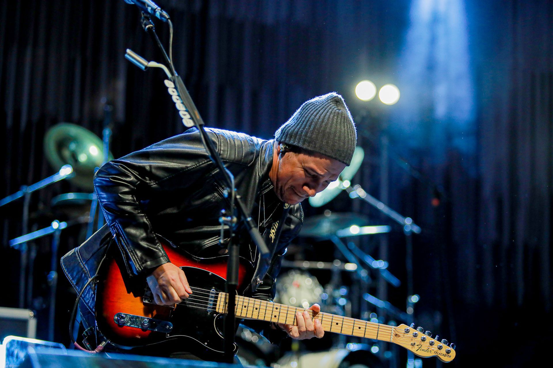 Um músico de gorro e jaqueta de couro toca uma guitarra elétrica sunburst em um palco com bateria e luzes desfocadas.
