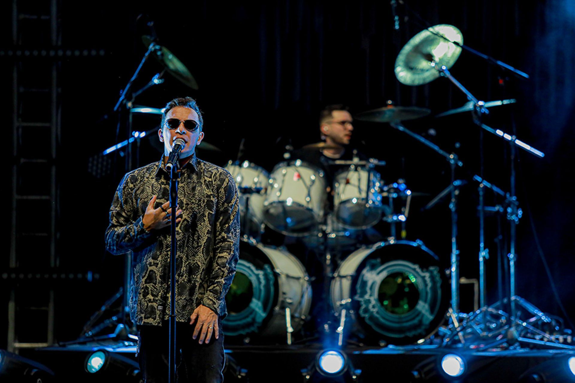 Um cantor com camisa estampada e óculos escuros se apresenta ao microfone, com um baterista tocando bateria atrás dela.