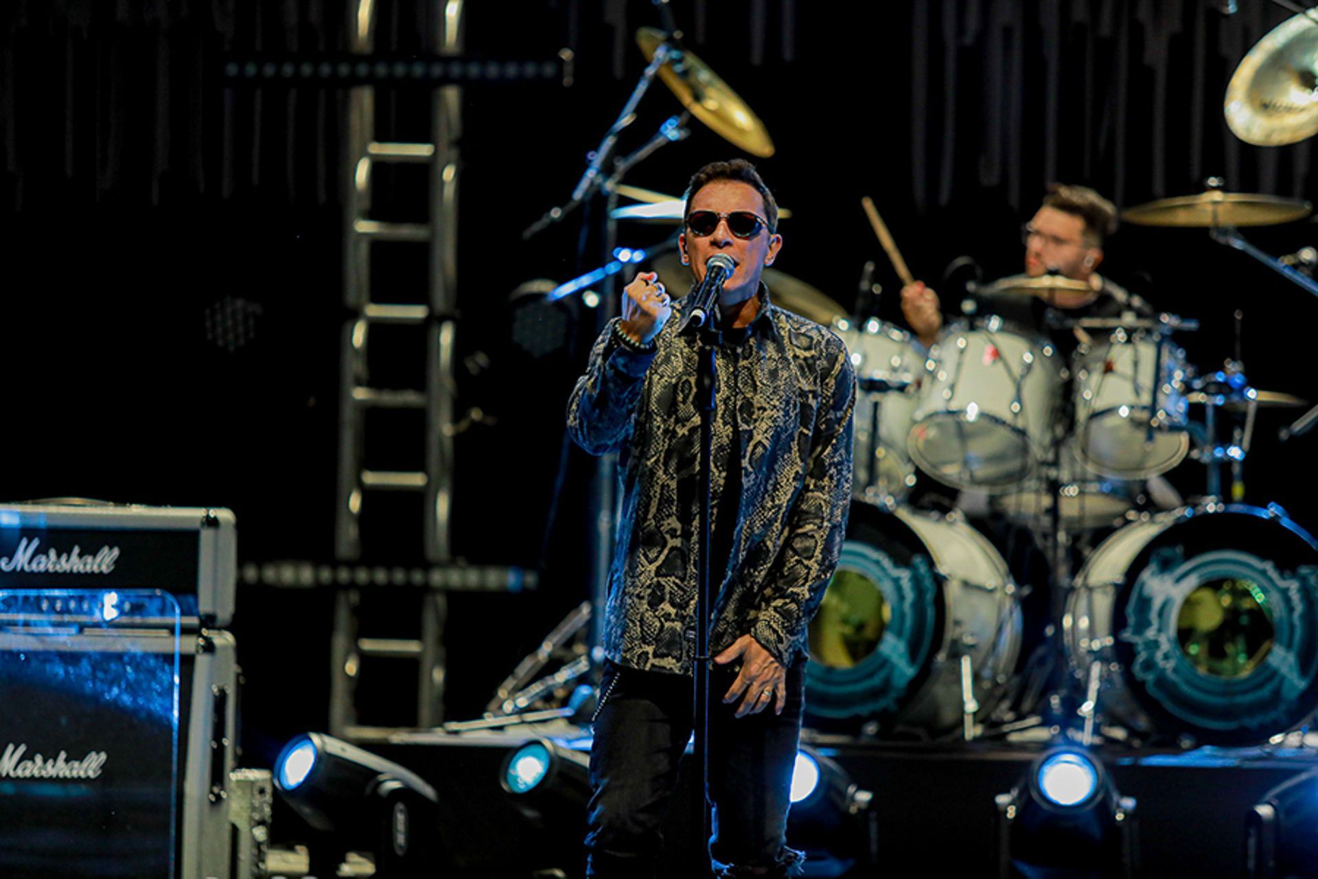 Um cantor de óculos escuros e camisa estampada se apresenta no palco com um baterista ao fundo.