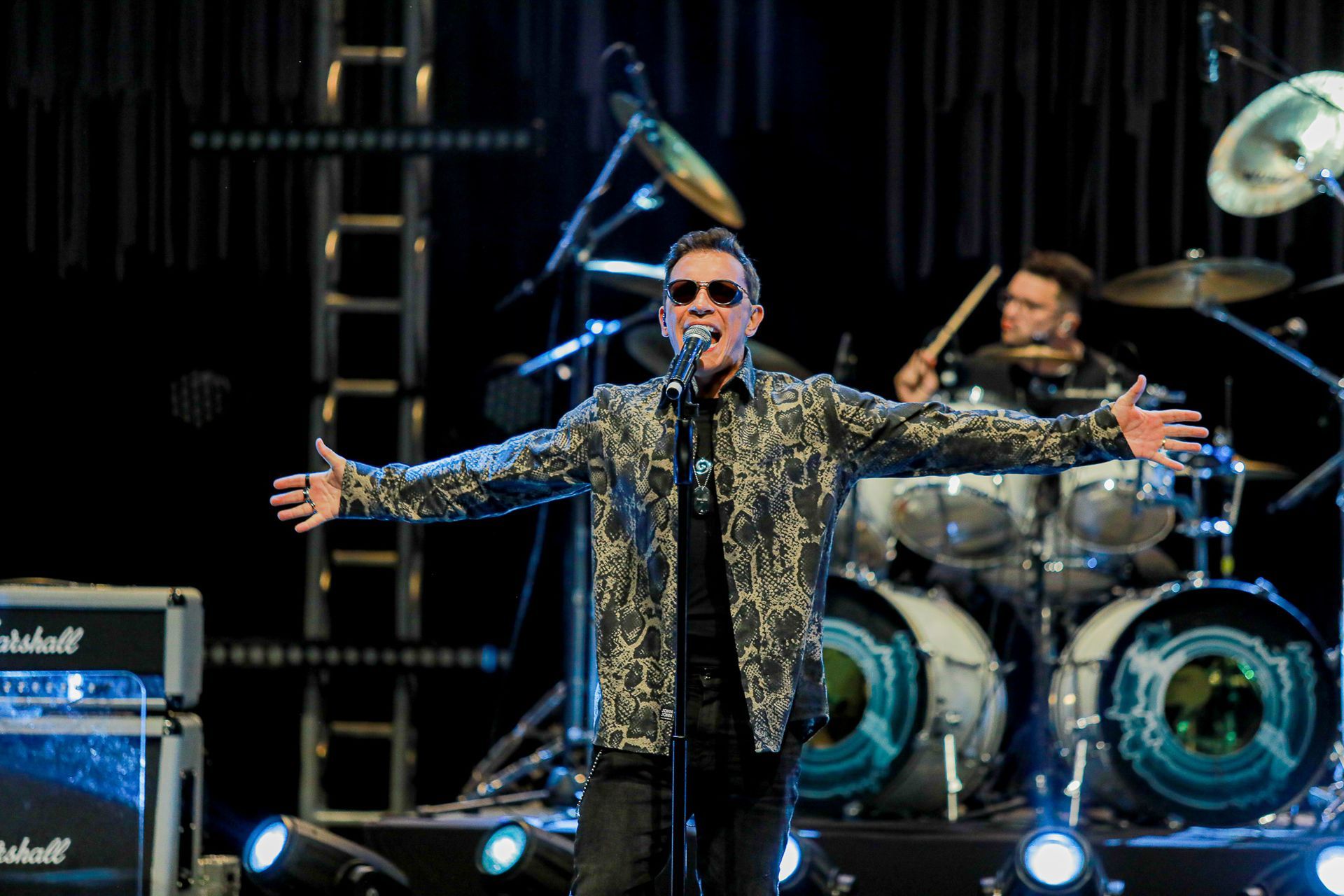 Um cantor de óculos escuros e jaqueta estampada se apresenta no palco com os braços estendidos; um baterista toca ao fundo.