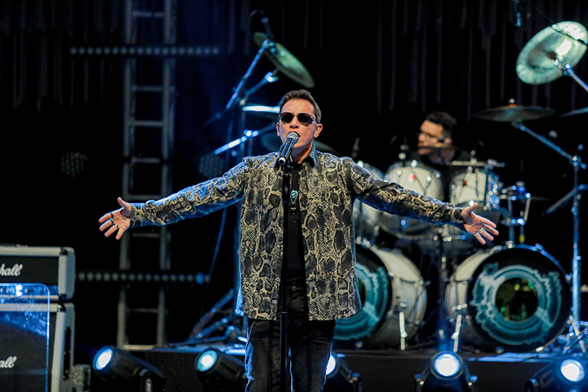 O vocalista principal, vestindo uma camisa estampada de botões e óculos escuros, se apresenta no palco de um show.