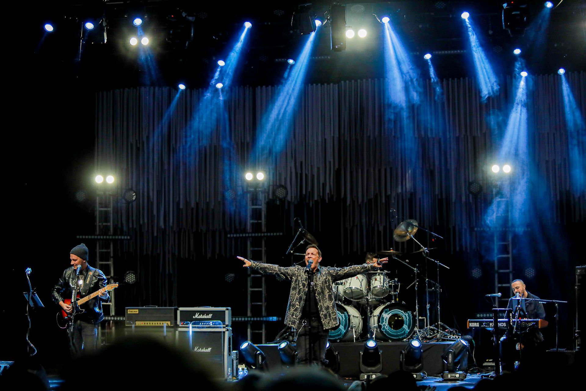Uma banda de rock se apresenta em um palco de shows com holofotes azuis iluminando os músicos contra um fundo escuro.