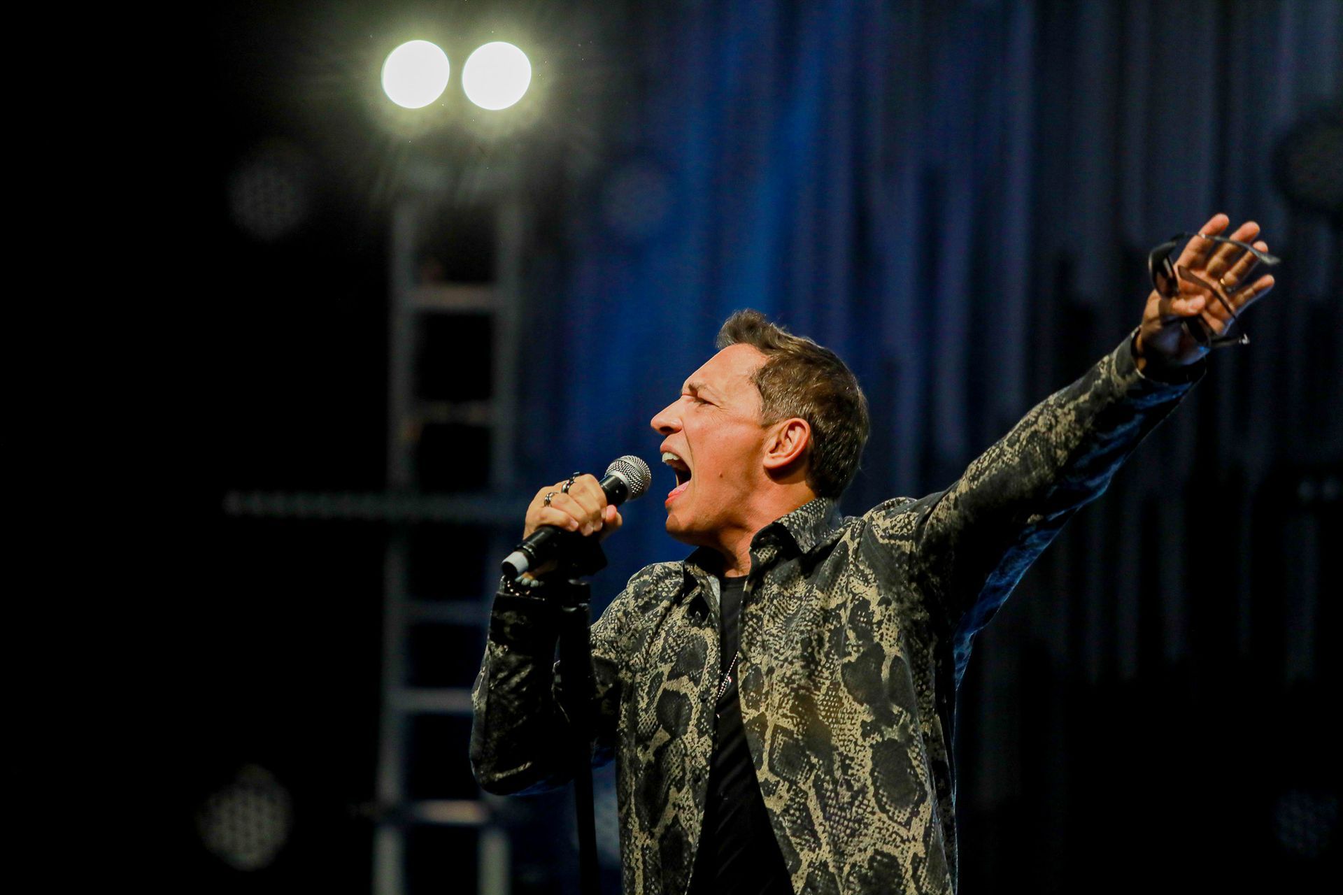 Um homem cantando em um microfone no palco, vestindo uma camisa escura estampada, com um braço erguido em direção à plateia.