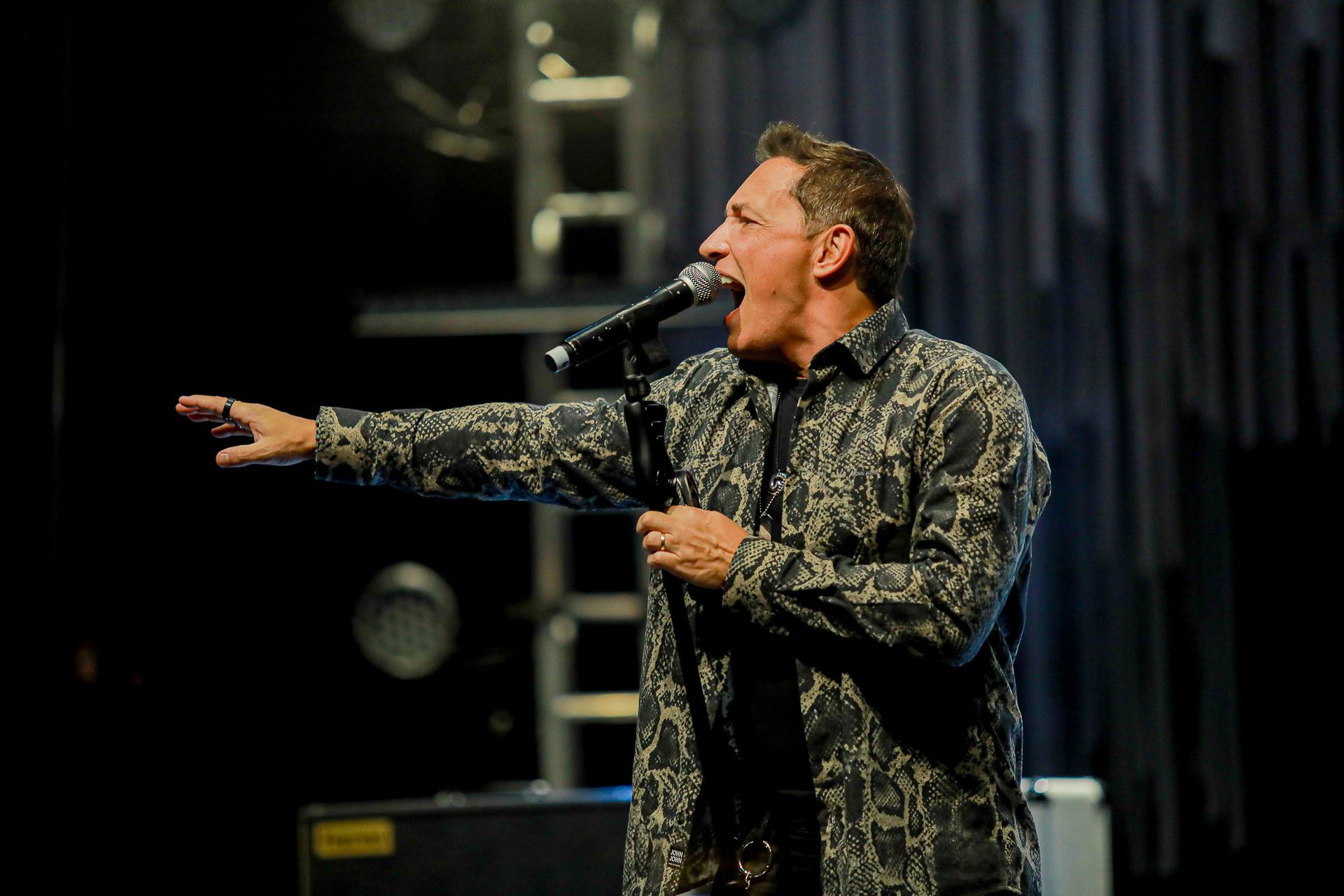 Um artista com um microfone na mão, cantando com paixão no palco enquanto gesticula com o braço direito.