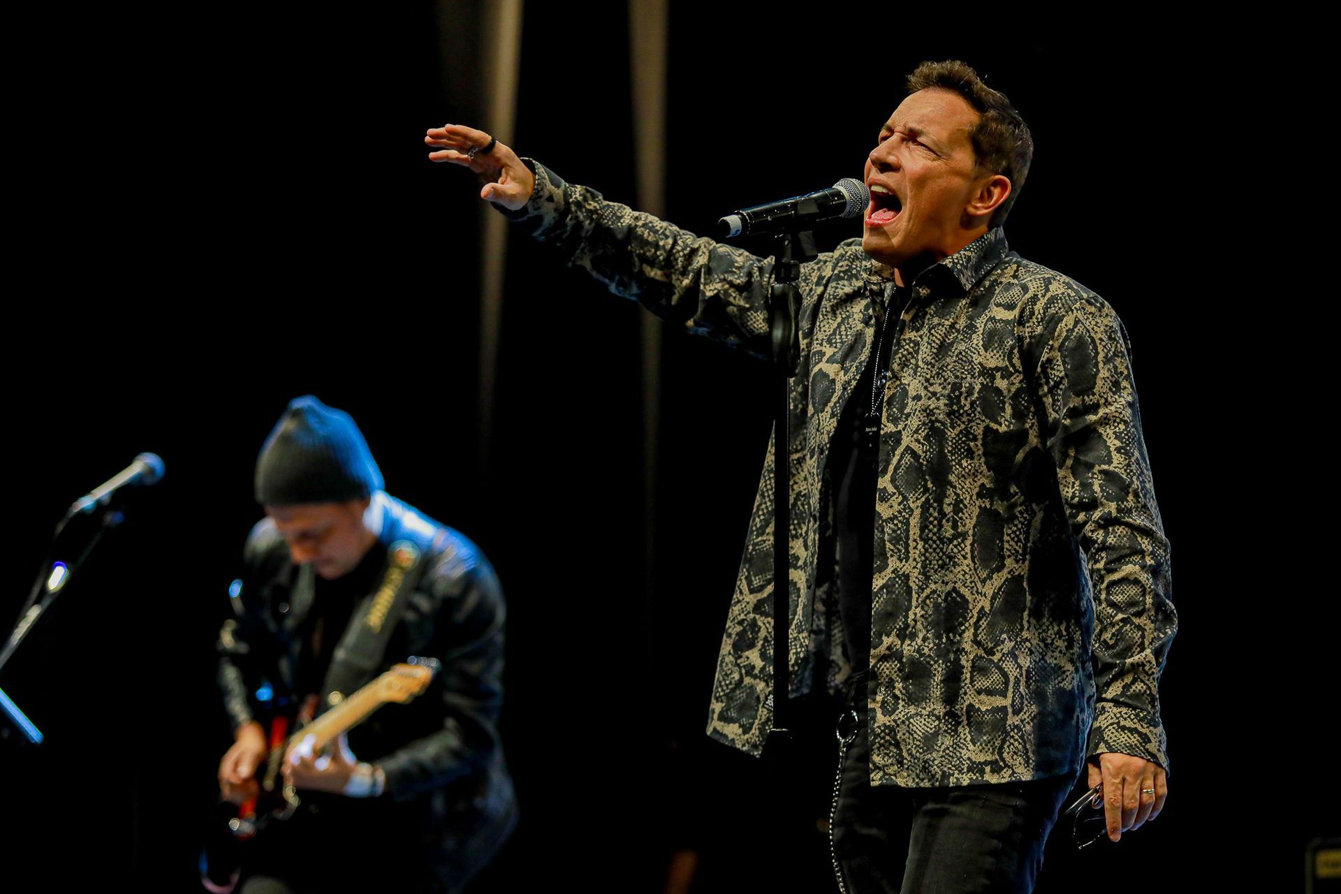 Um cantor vestindo uma camisa estampada de botões se apresenta em um palco escuro, enquanto um guitarrista toca.
