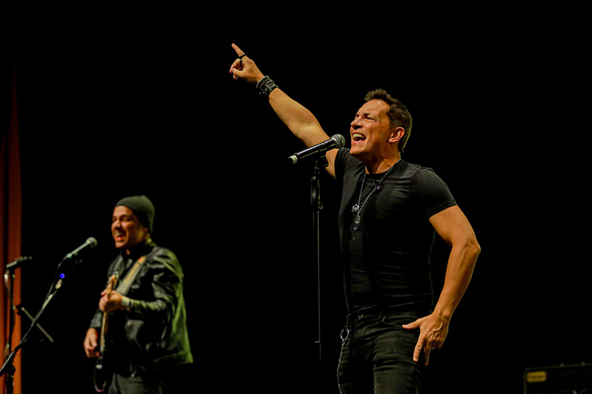 Um cantor apontando para cima enquanto se apresenta no palco com um guitarrista atrás dele, em um fundo escuro.