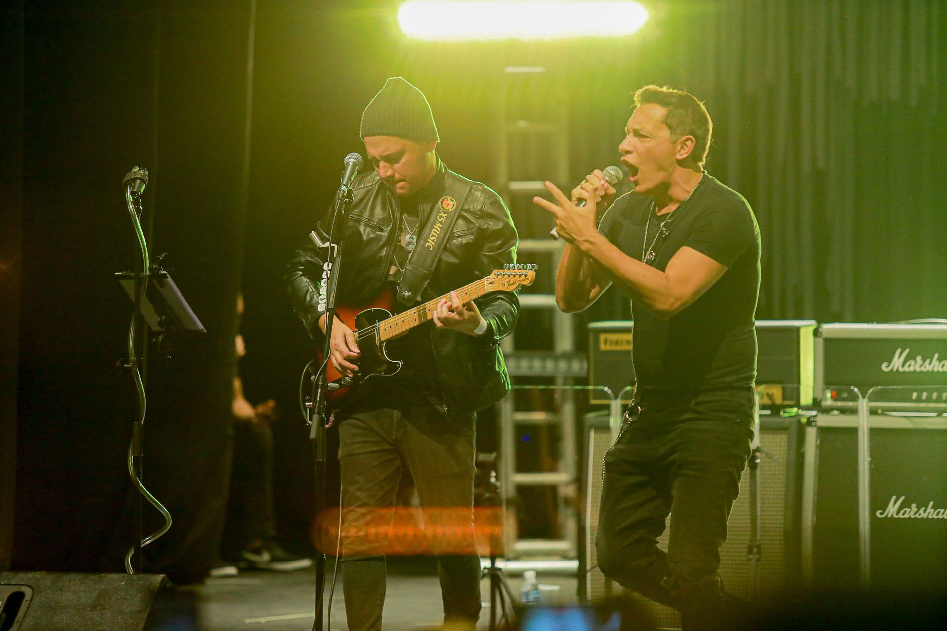 Dois artistas no palco sob iluminação amarela: um tocando guitarra elétrica, o outro cantando ao microfone.