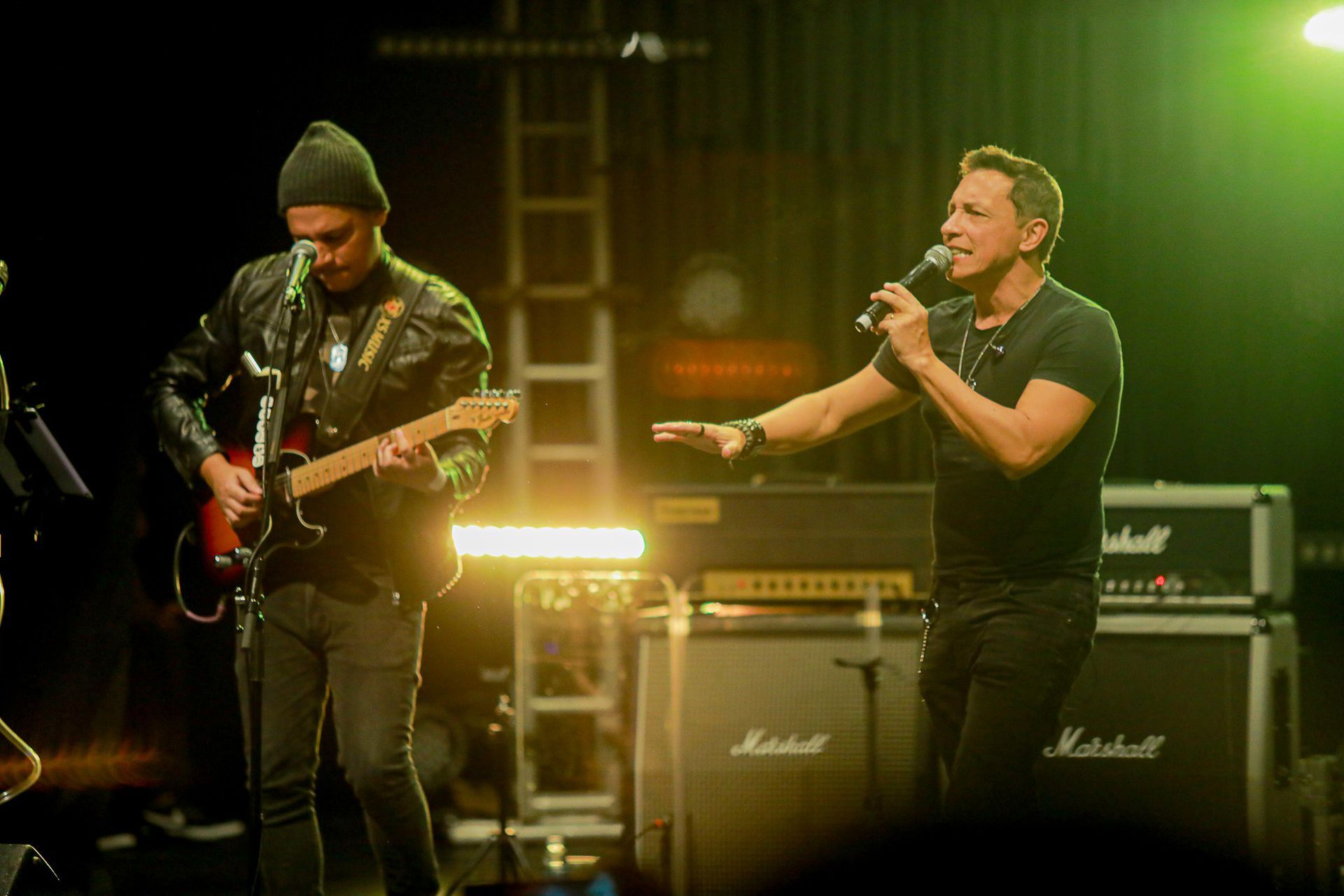 Dois homens se apresentando no palco; um cantando ao microfone, o outro tocando guitarra elétrica.