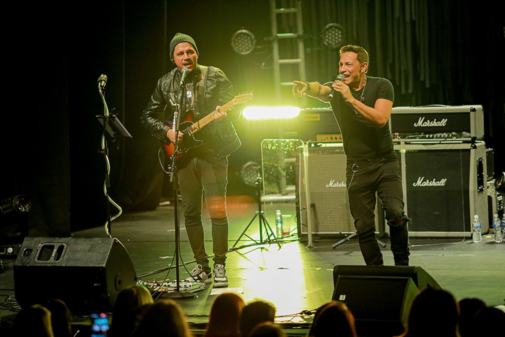 Dois artistas em um palco pouco iluminado: um tocando guitarra, o outro cantando em um microfone e apontando para a frente.