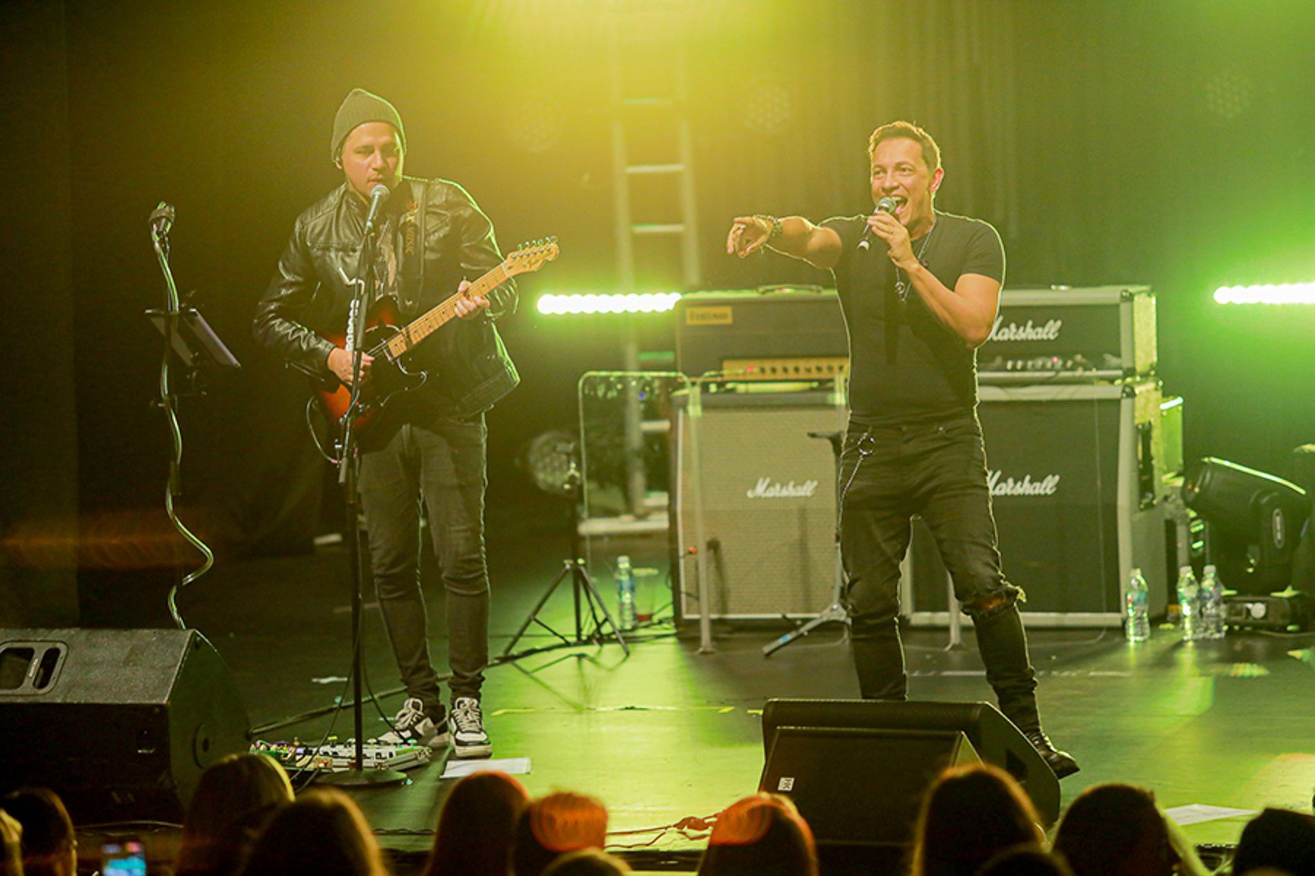Dois músicos se apresentam em um palco com amplificadores Marshall, um tocando guitarra e o outro cantando sob luzes verdes.