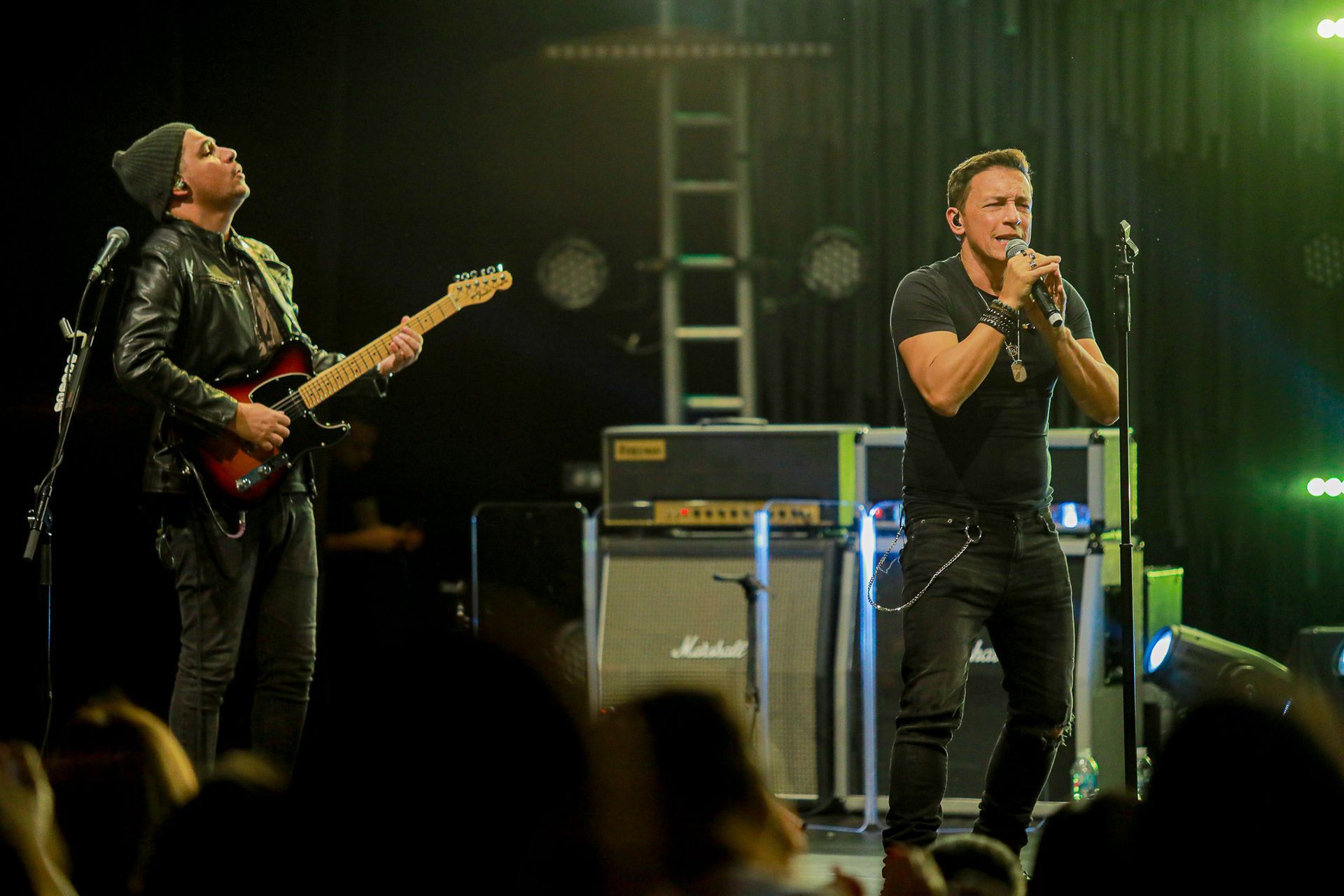 Um músico toca guitarra elétrica enquanto outro canta ao microfone em um palco diante de uma plateia.