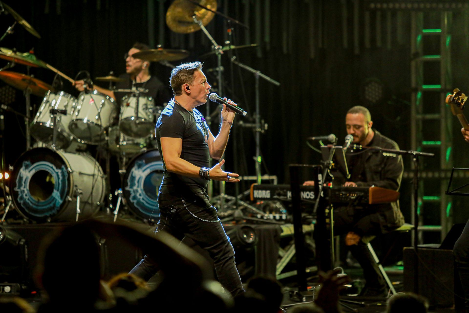 Uma banda se apresenta no palco com um vocalista ao microfone, um baterista ao fundo e um tecladista à direita.